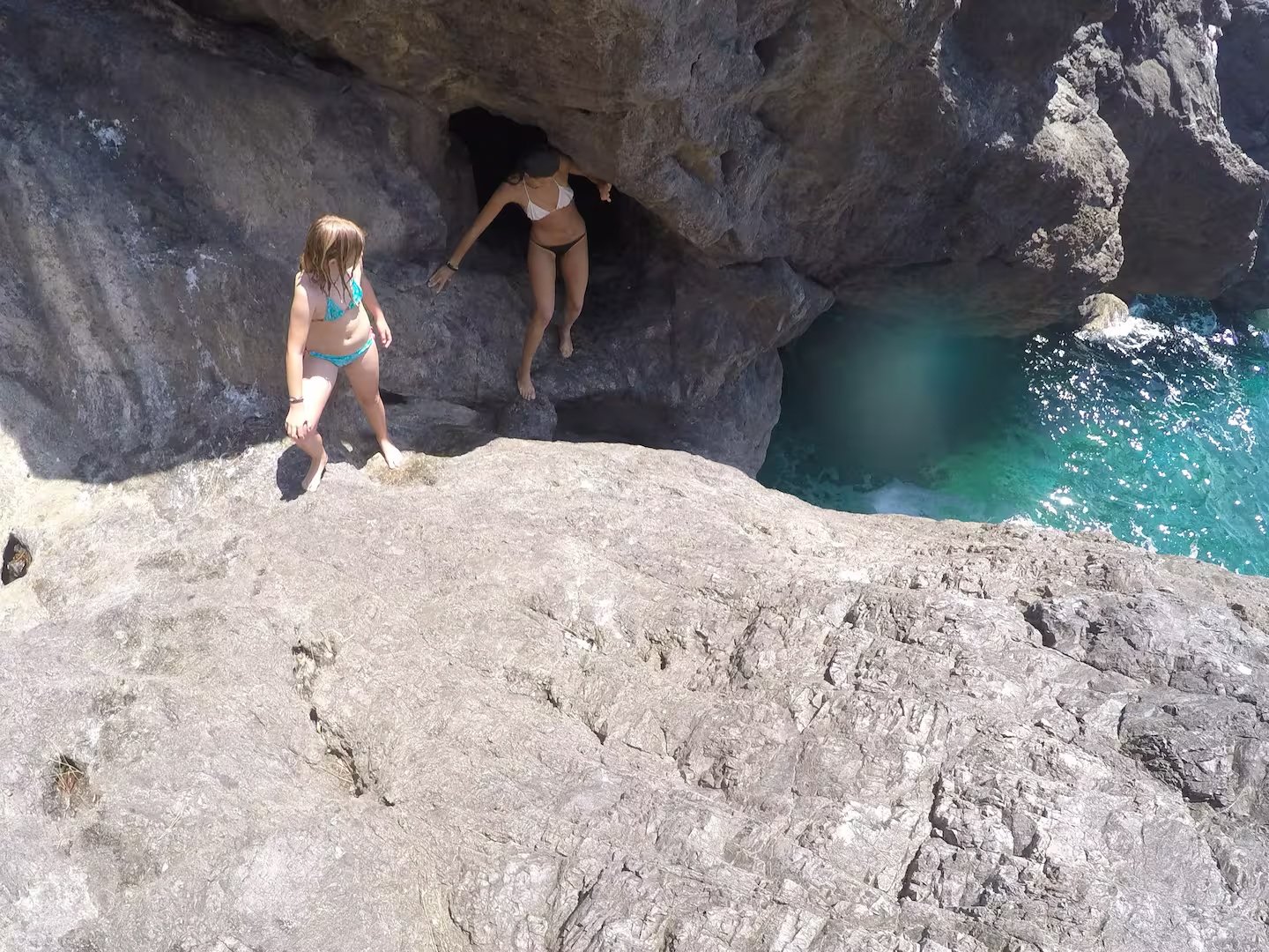 Two people in swimsuits exploring rocky cliffs by the ocean with a cave opening.