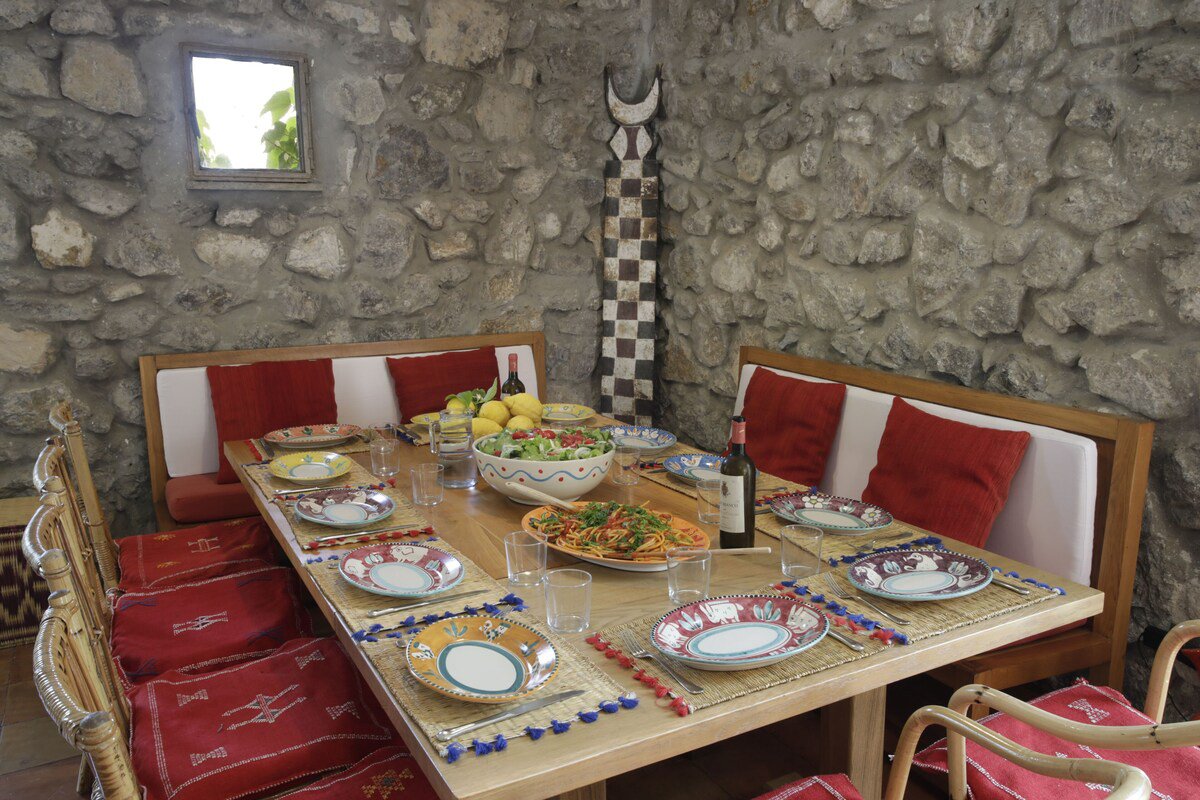 Dining table set with colorful plates, a bowl of salad, a platter of pasta, a bottle of wine, and fruit, in a room with a stone wall and red cushions.