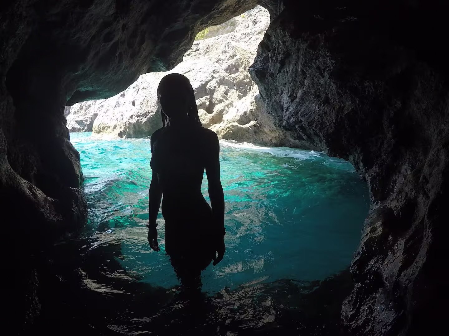 Silhouette of a person standing in a rocky cave entrance with turquoise water in the background.