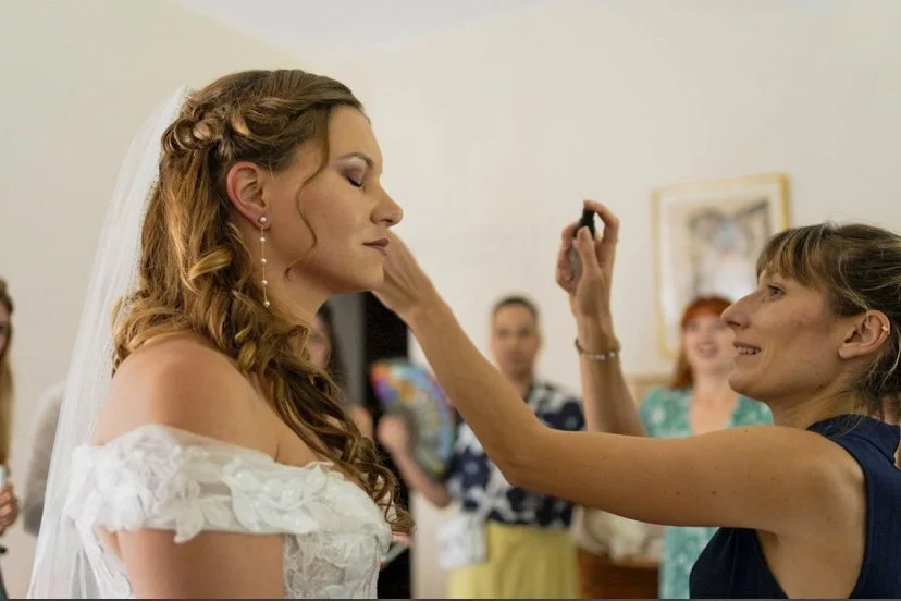 A bride with long wavy hair and a veil, getting her makeup or powder done by a woman in a navy dress, at a wedding reception with guests in the background.