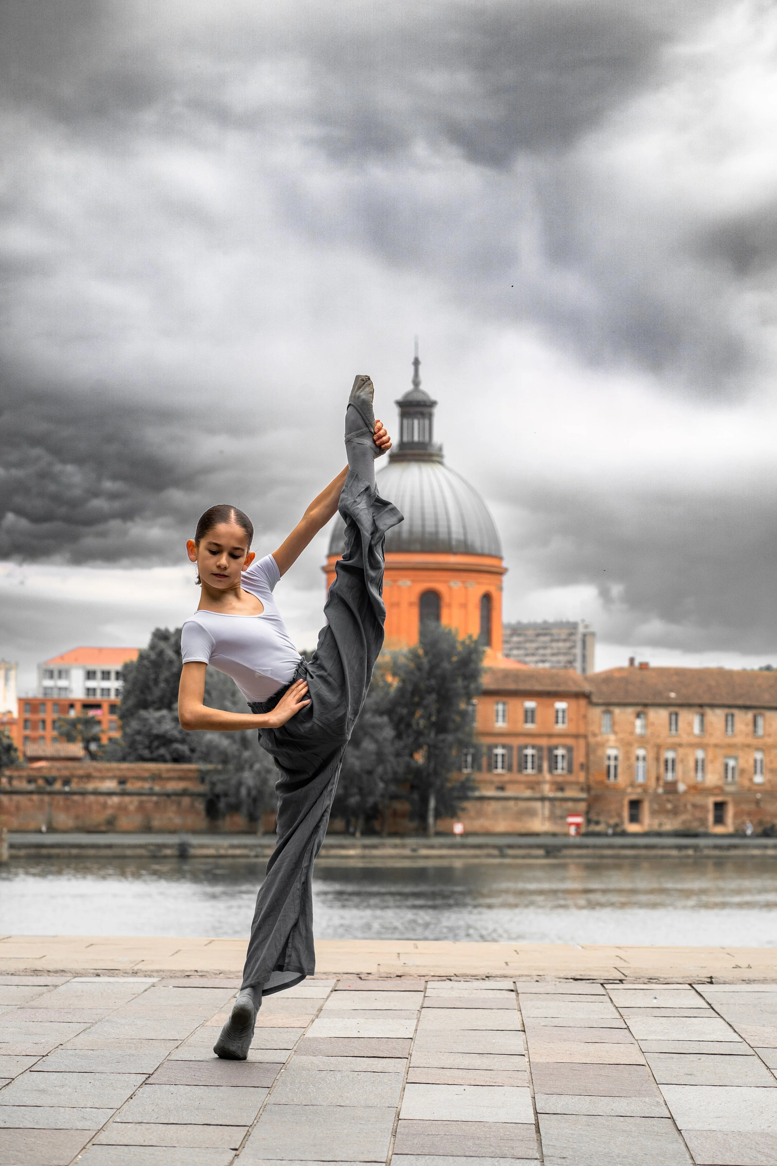 Young female dancer holding a high leg position outdoors on a paved riverfront promenade with a cityscape, including a domed building, in the background under dark cloudy sky.