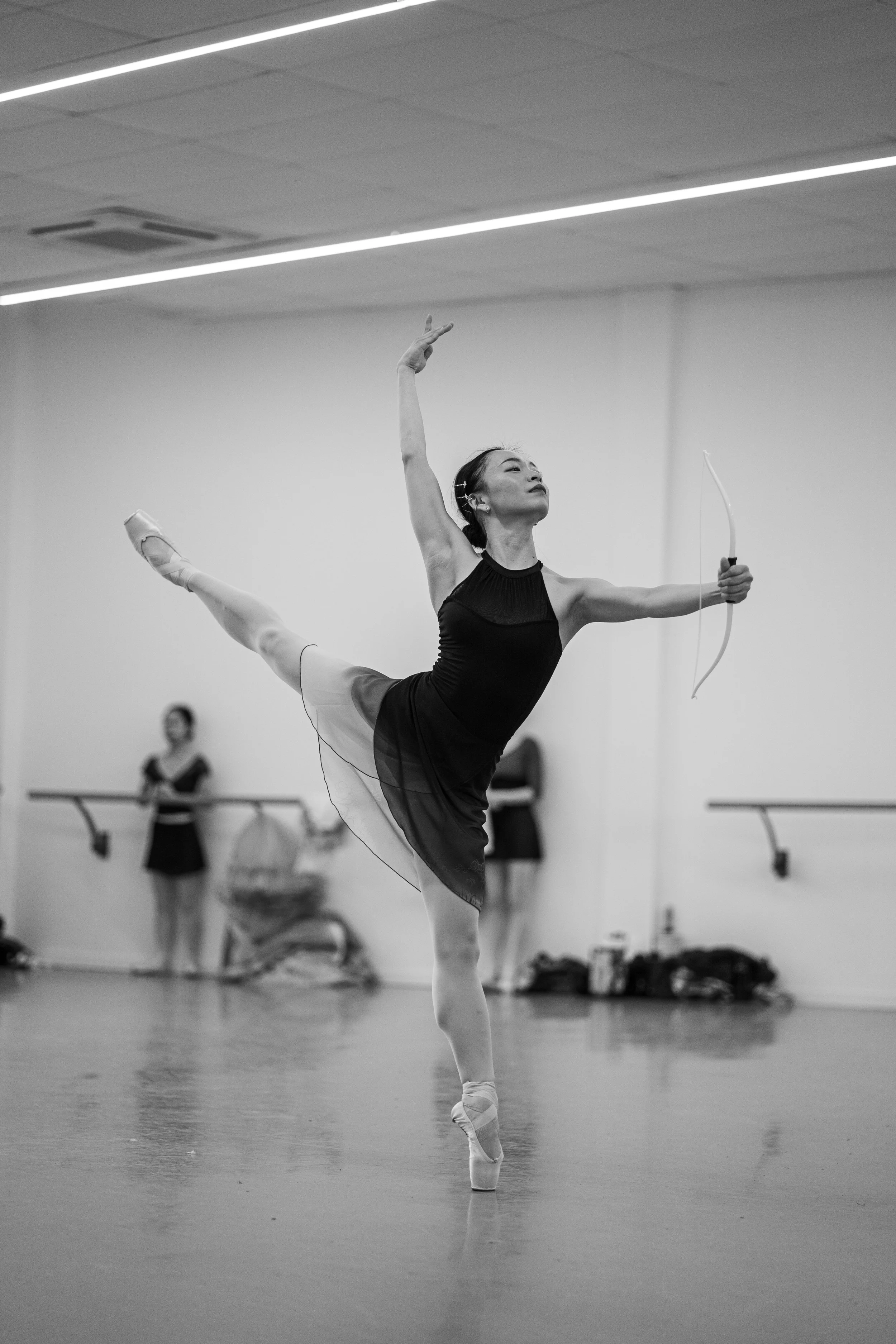 A ballet dancer practicing in a studio, balancing on toes while holding a bow with an arrow, wearing a black dress and ballet slippers.