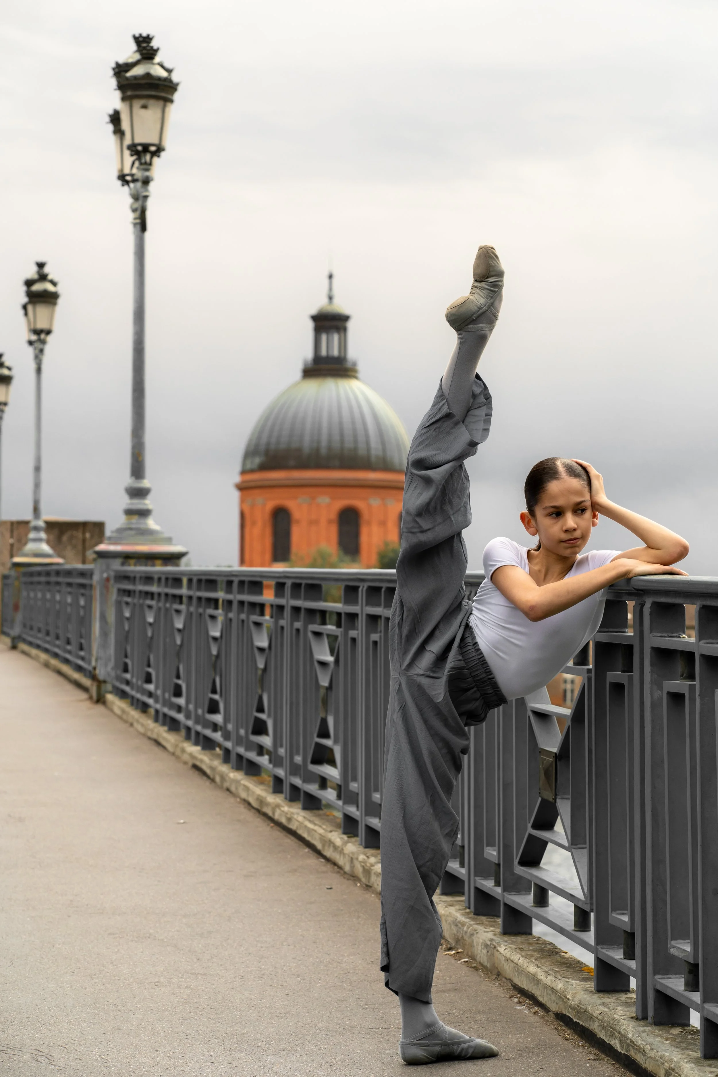 A young girl practicing a high leg ballet stretch on a bridge with city buildings and a domed church in the background, overcast sky.