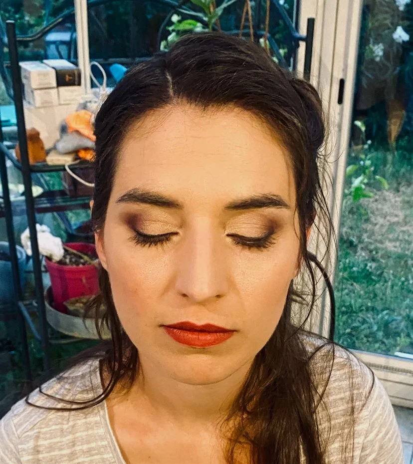 A woman with dark hair, closed eyes, and makeup, sitting indoors near a window with a garden view in the background.