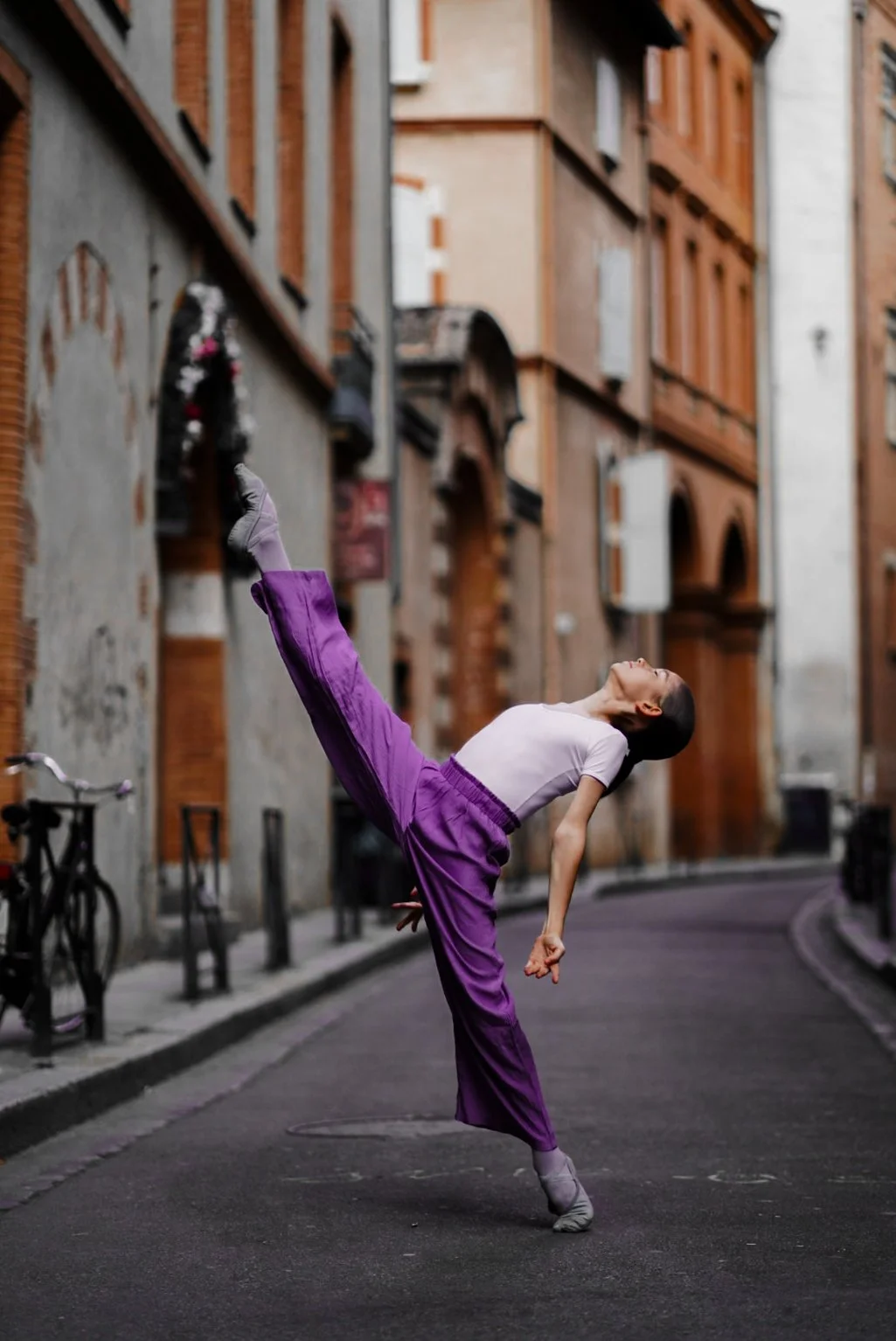A woman performing a ballet pose on a city street in front of red brick buildings, wearing purple pants and a white shirt.