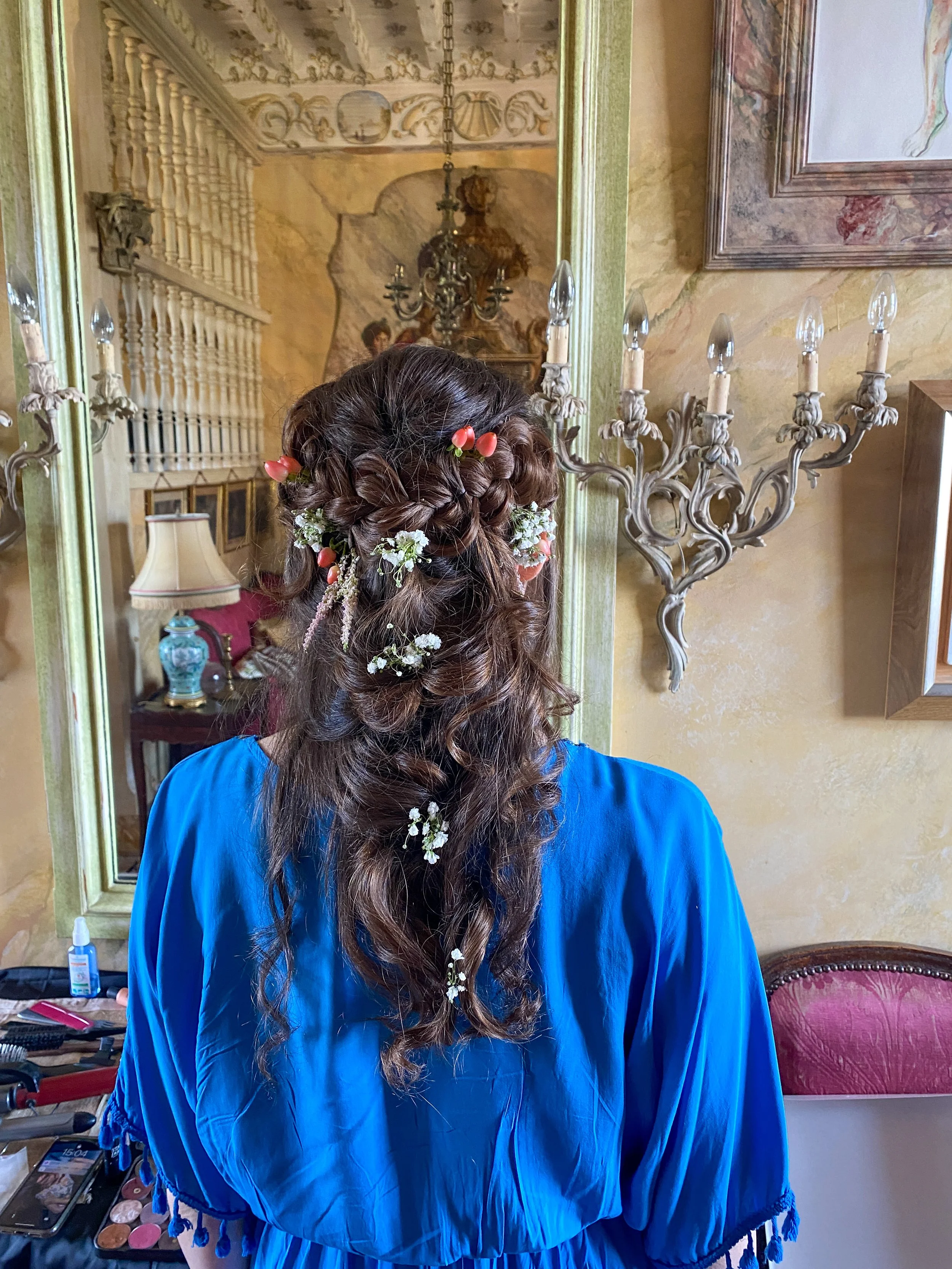 Back of a woman with long, curly brown hair decorated with small white flowers and pink buds, wearing a bright blue dress, in front of a mirror in a vintage styled room.
