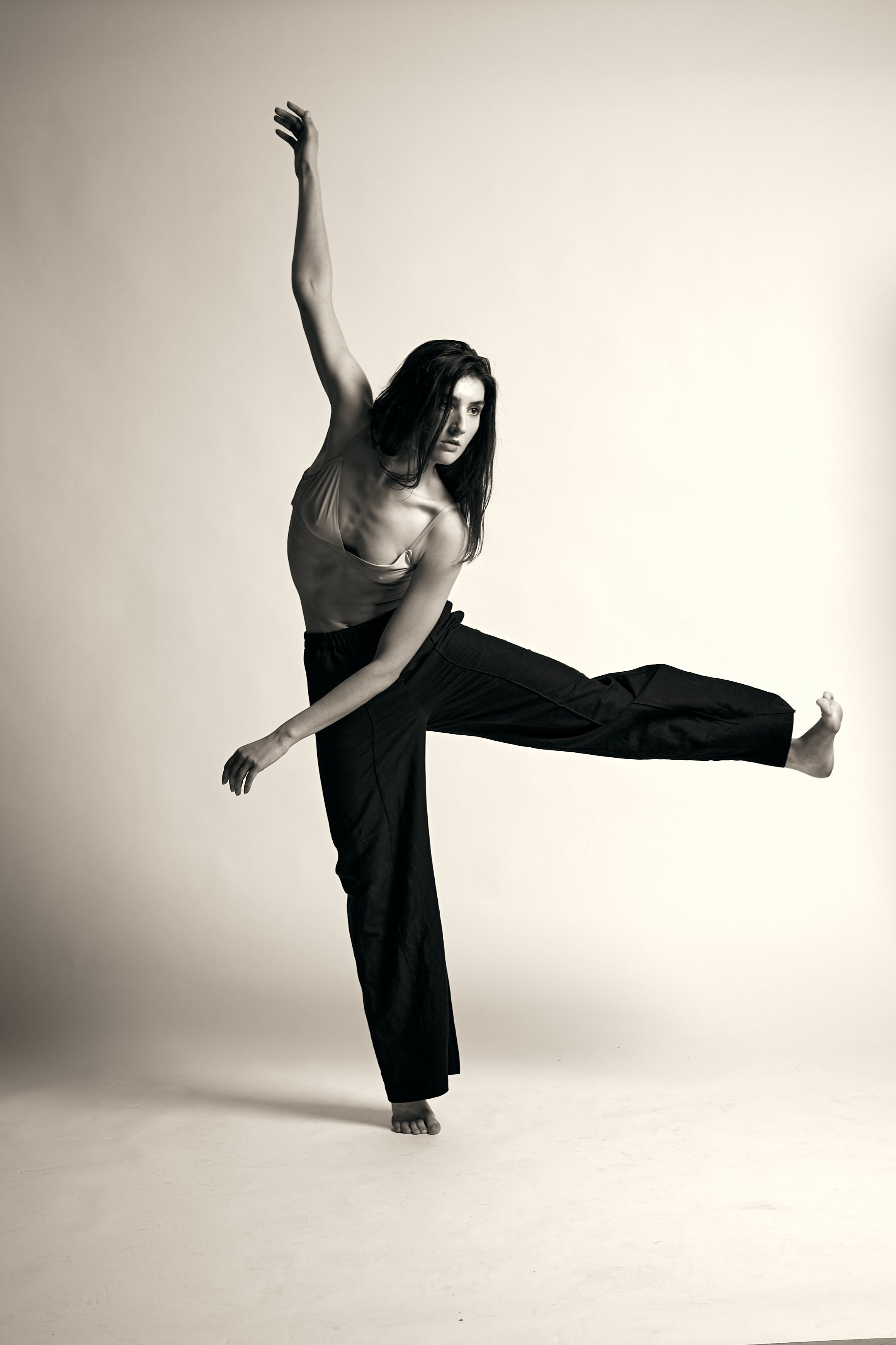 A woman dancing or performing a pose, standing on one foot with the other leg extended sideways and arms raised overhead, in a studio with a plain background.