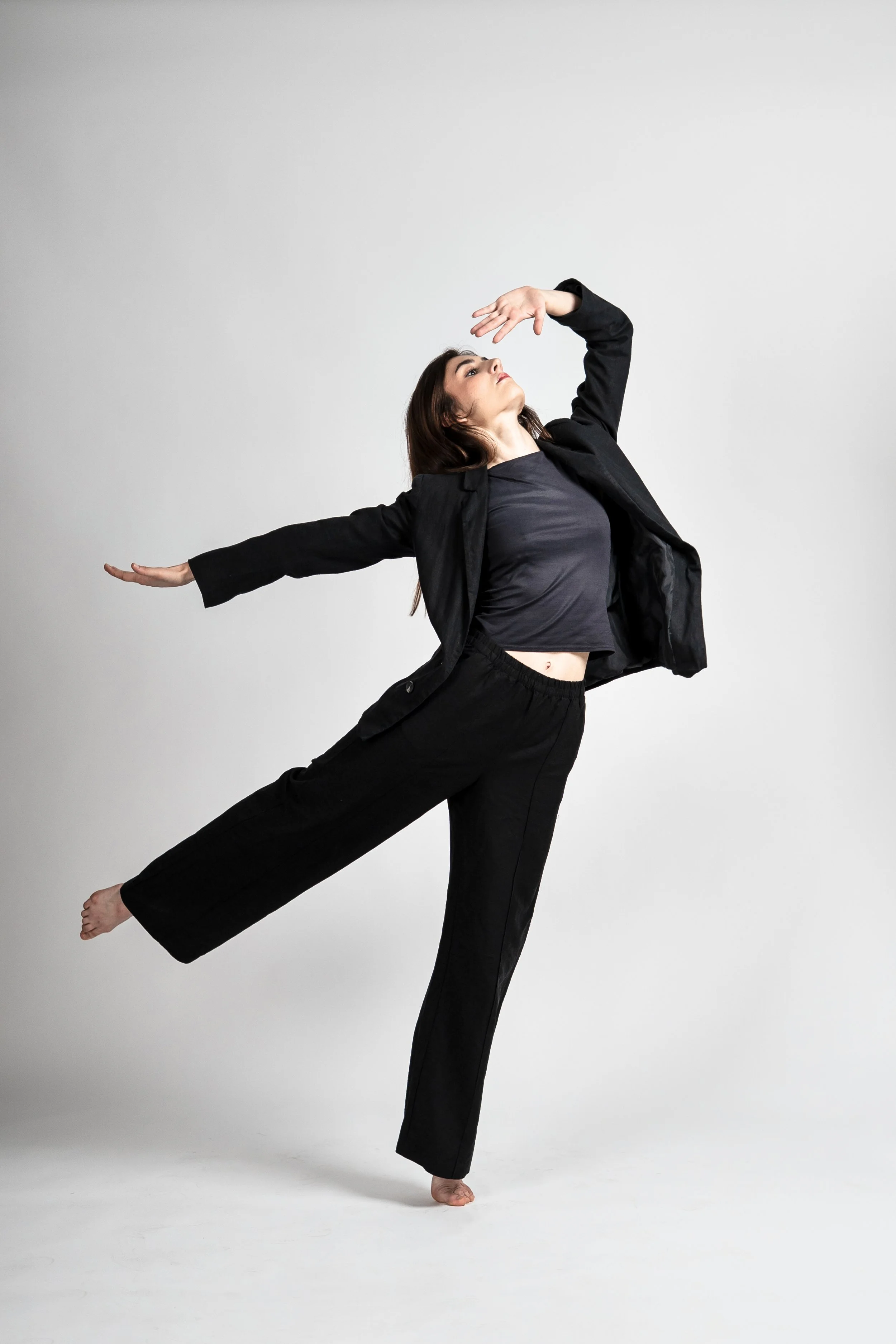 A woman in black pants, a dark t-shirt, and a blazer is balancing on one leg with arms outstretched, posing against a plain white background.