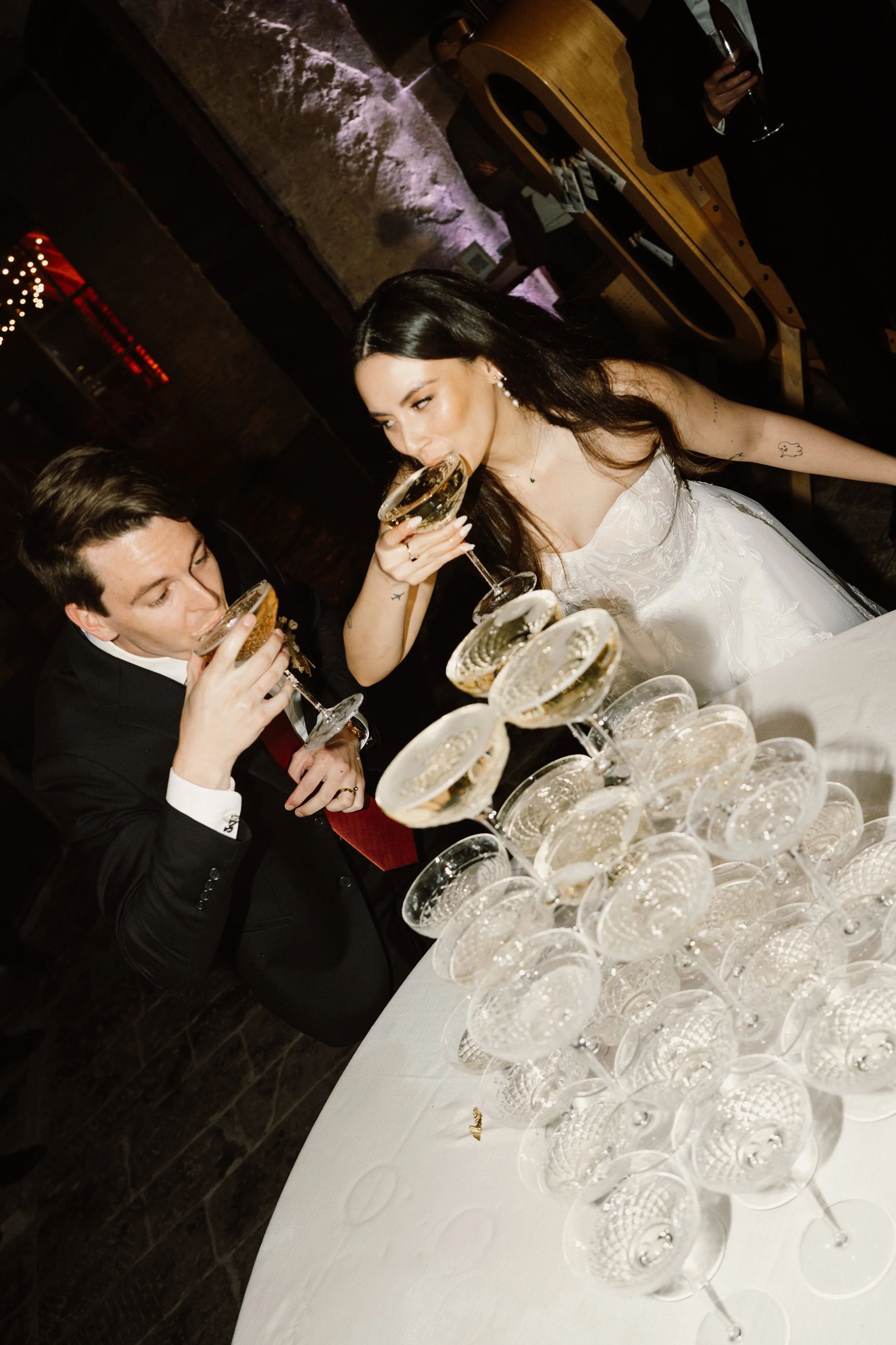 A bride and groom toast with champagne at a wedding reception, surrounded by stacked champagne glasses on a table.
