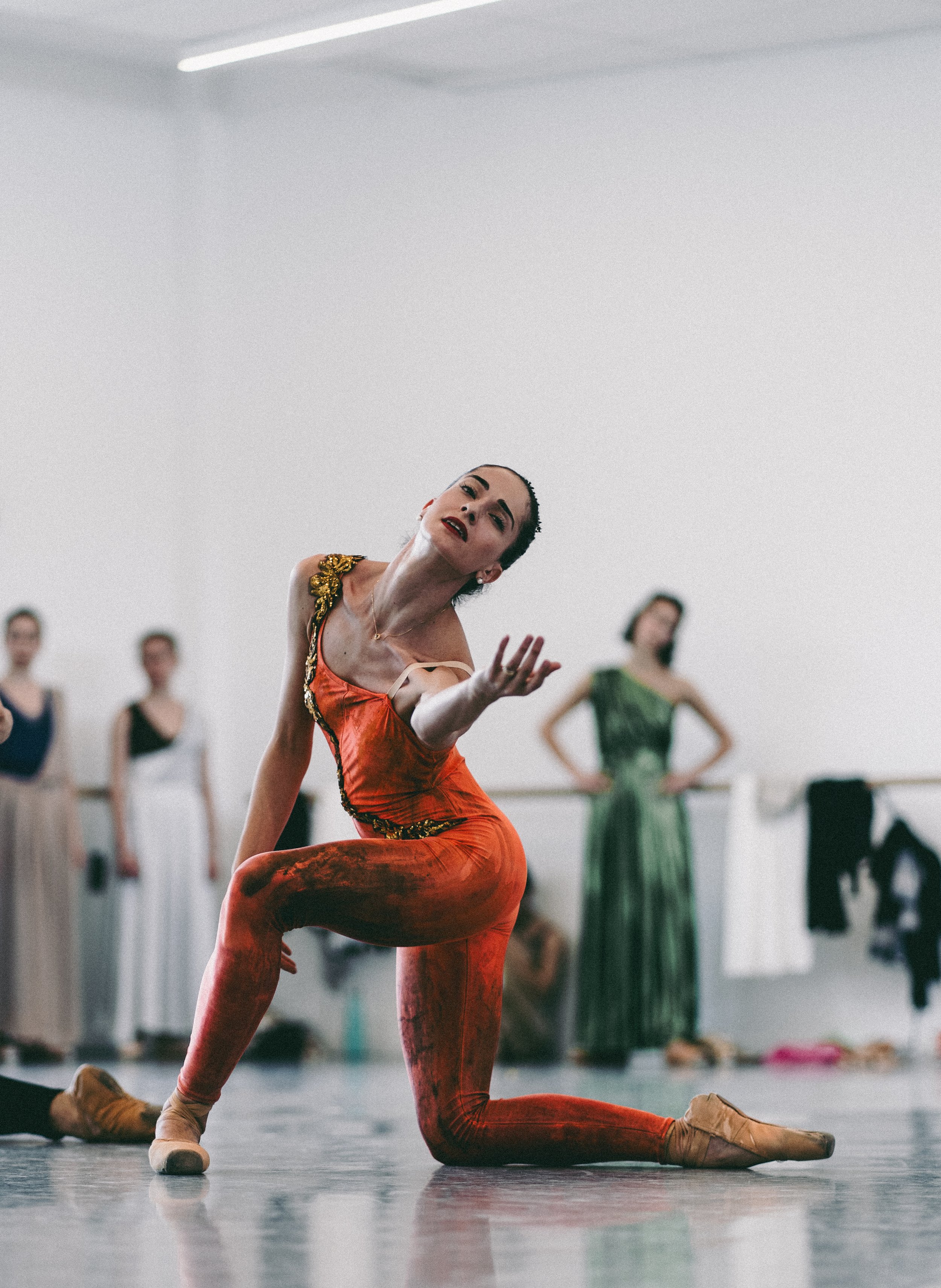 Ballet dancer in red costume performing a pose in a dance studio with other dancers in the background.