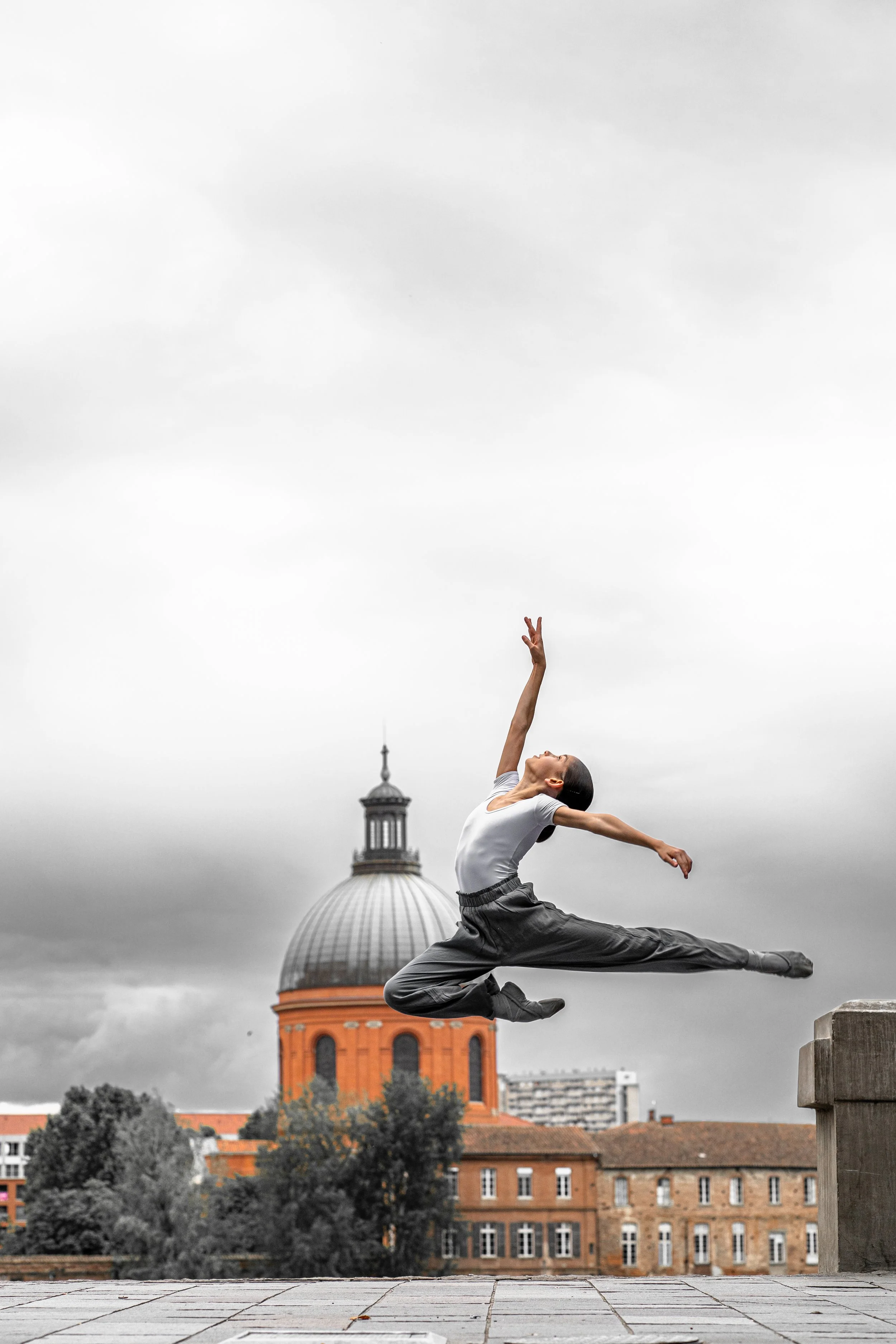 A dancer performing a leap in midair outdoors with a domed building and cloudy sky in the background.