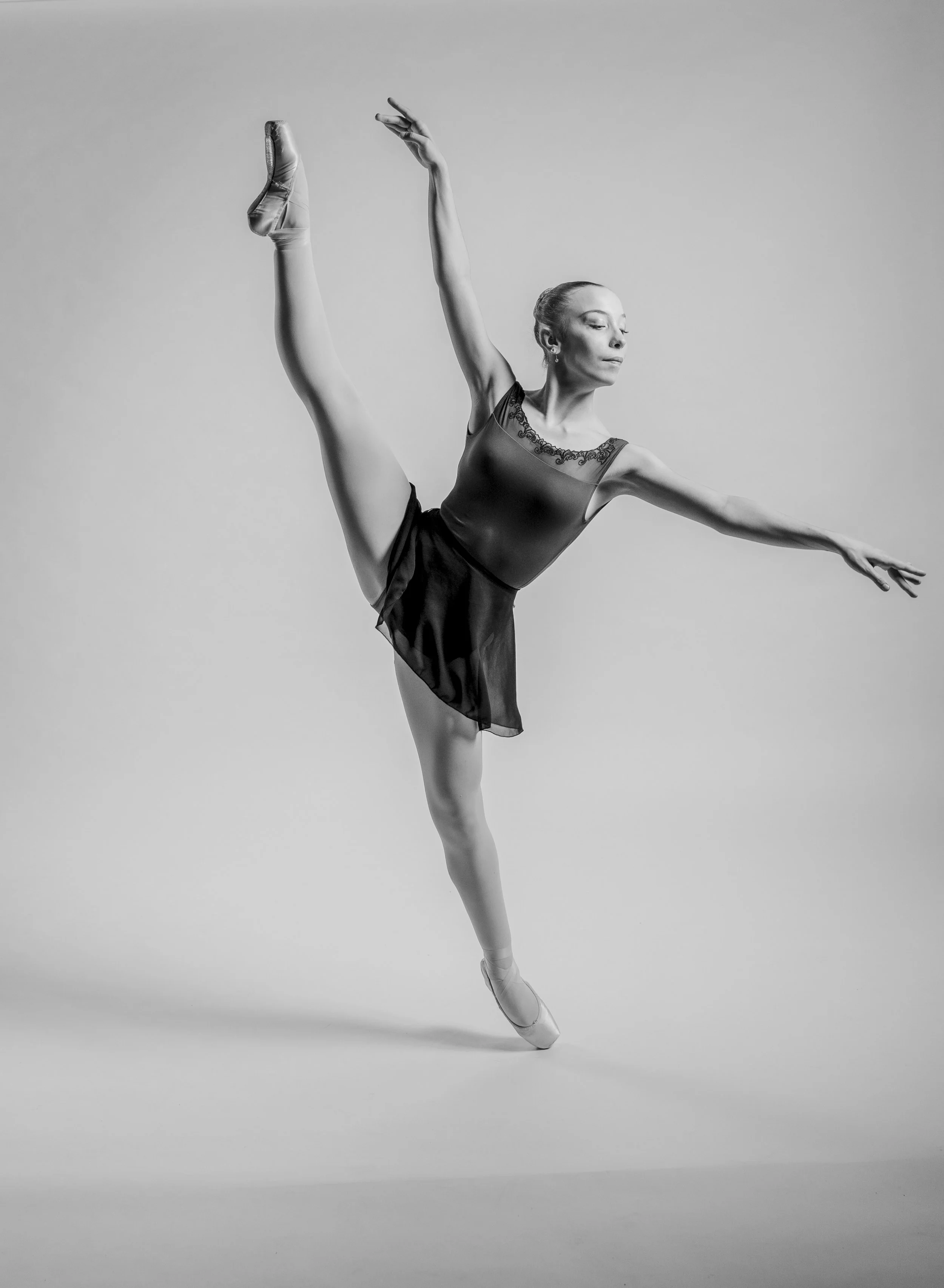 A ballet dancer in a black dress and pointe shoes performing an arabesque pose against a plain background.