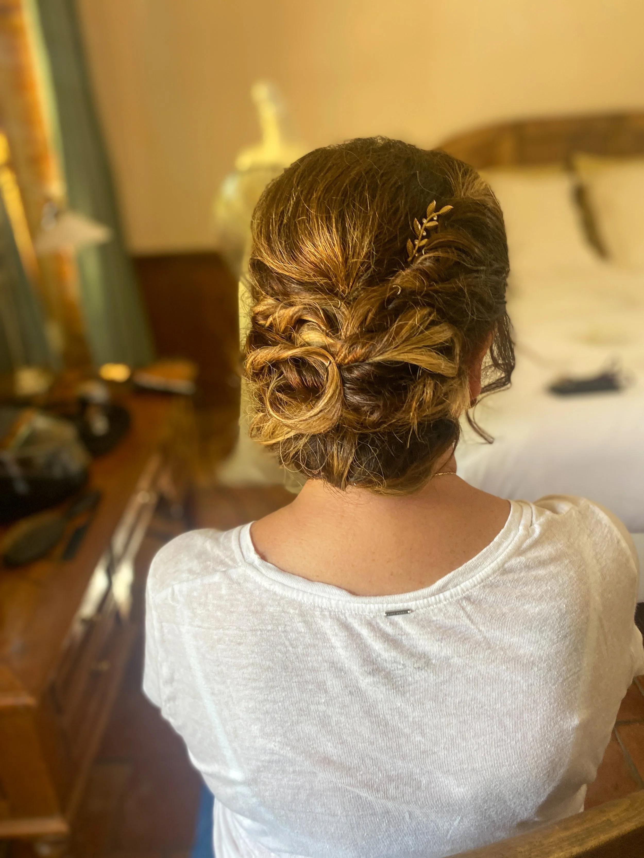 Back view of a woman with intricately styled hair in an updo, decorated with a small gold leaf hair accessory, wearing a cream-colored top, in a warmly lit room with a bed and wooden furniture.