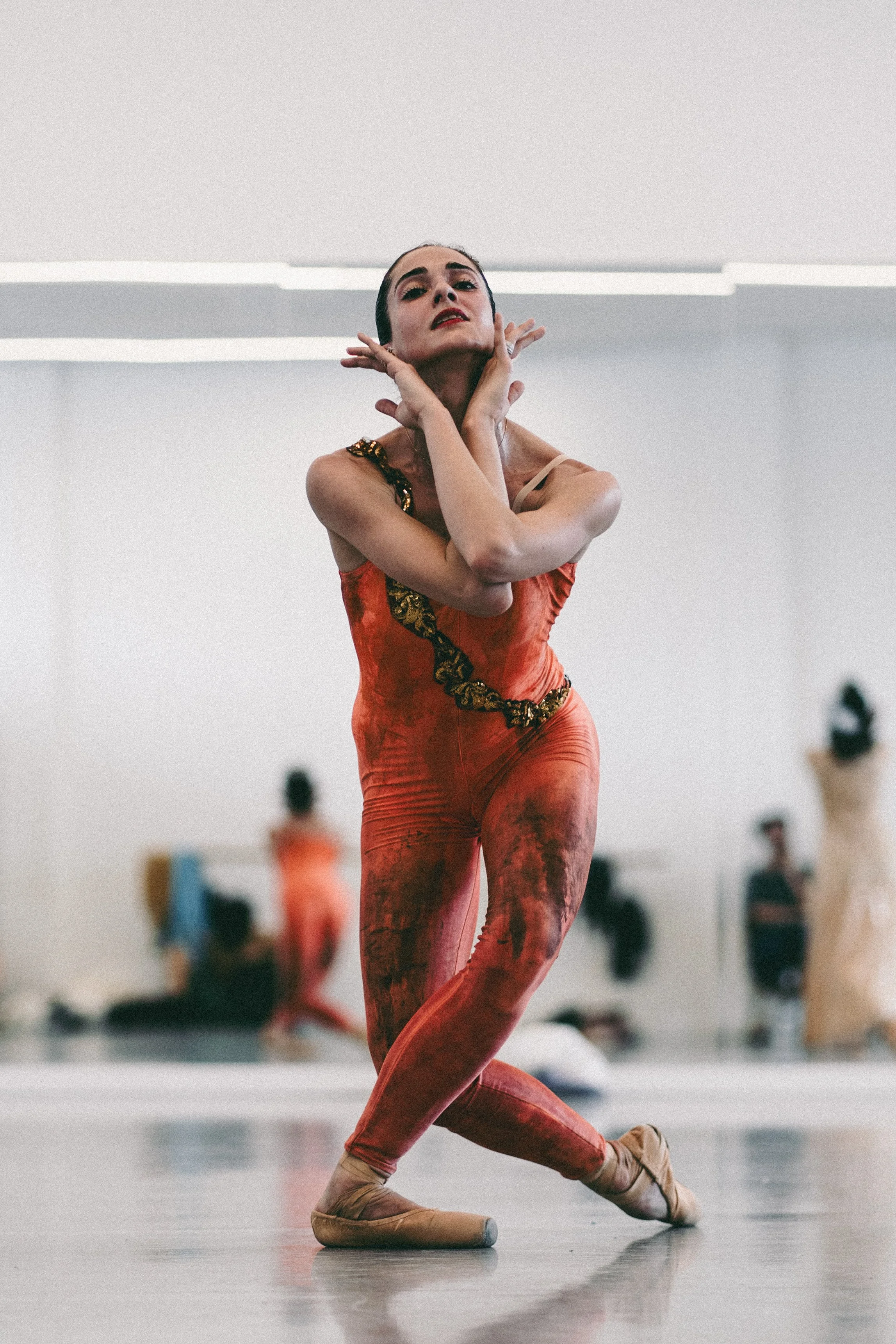 A dancer performing in a ballet studio, wearing a vibrant orange costume with gold accents and positioned on a mirrored floor.
