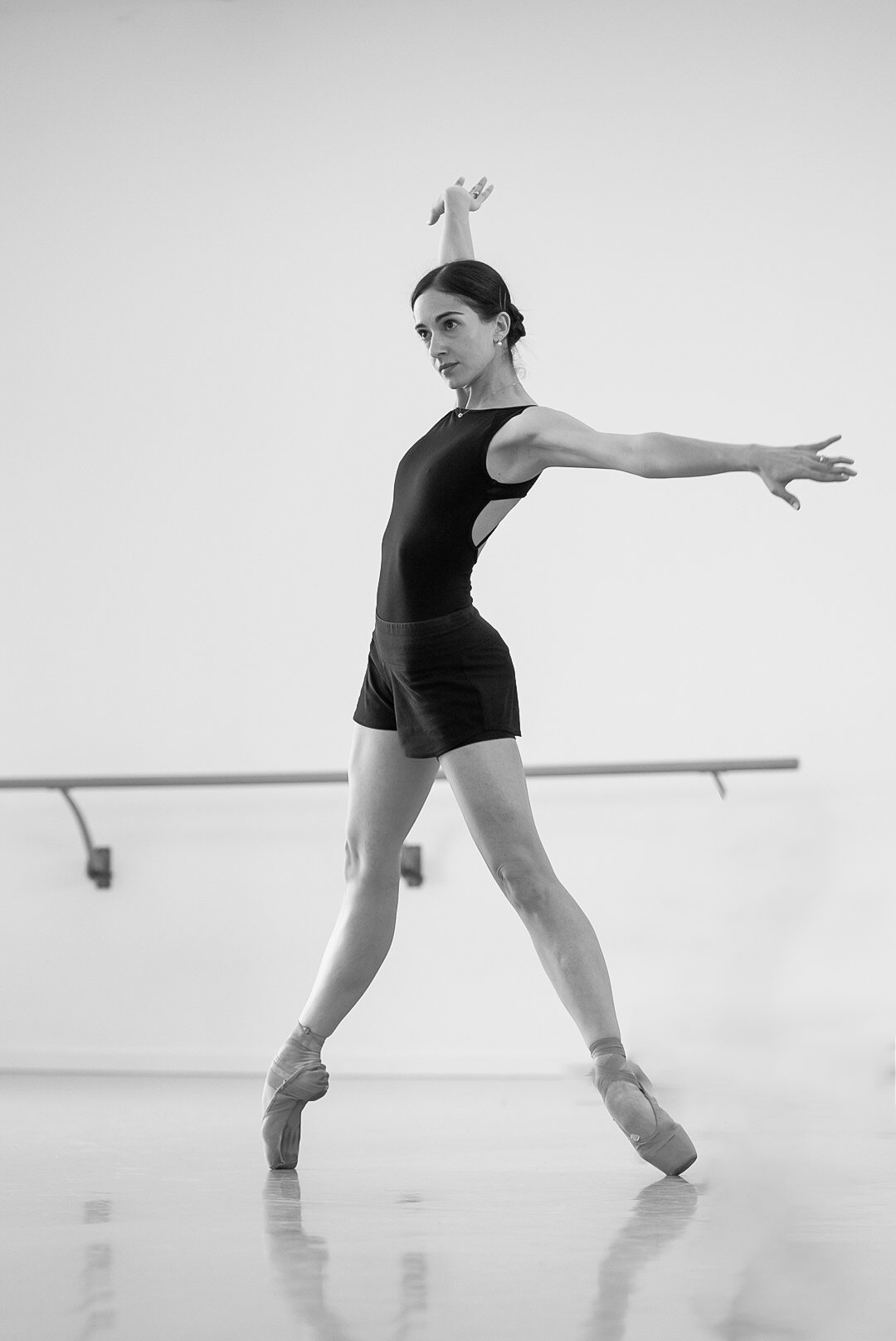 A female ballet dancer practicing in a studio, wearing a black leotard and ballet slippers, standing on her toes with arms extended.