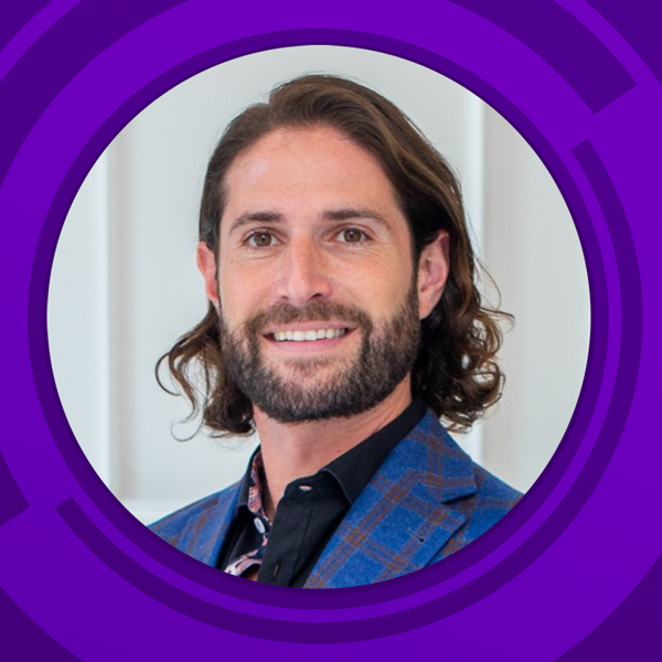 Headshot of a man with shoulder-length brown hair and a beard, smiling, wearing a blue plaid blazer and black shirt, standing in front of a white background with a purple circular frame around him.