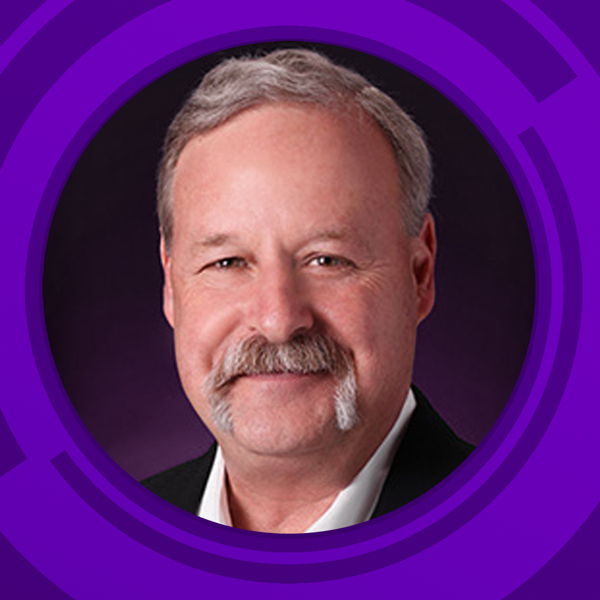 A middle-aged man with light skin, short gray hair, and a mustache, wearing a black suit and white shirt, smiling at the camera against a purple patterned background.