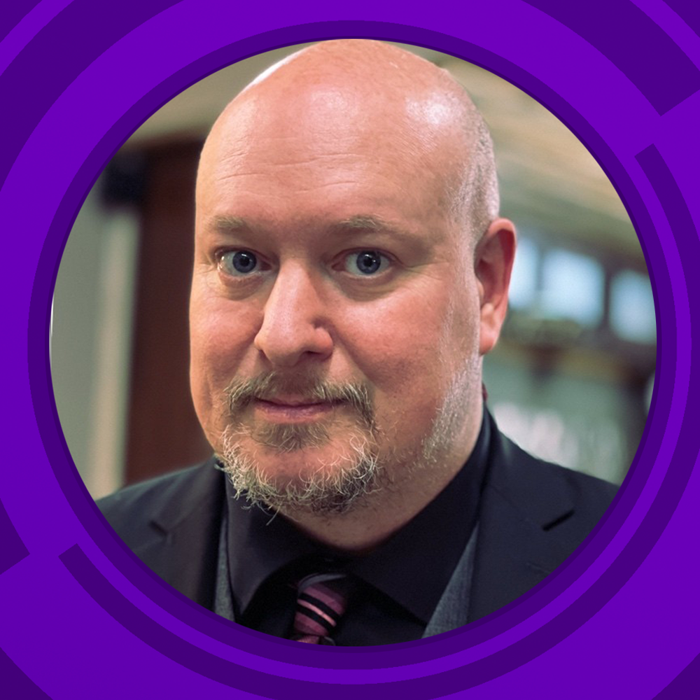 A close-up portrait of a bald man with blue eyes, a beard, and a mustache, wearing a black suit, shirt, and a purple striped tie, with a blurred indoor background.