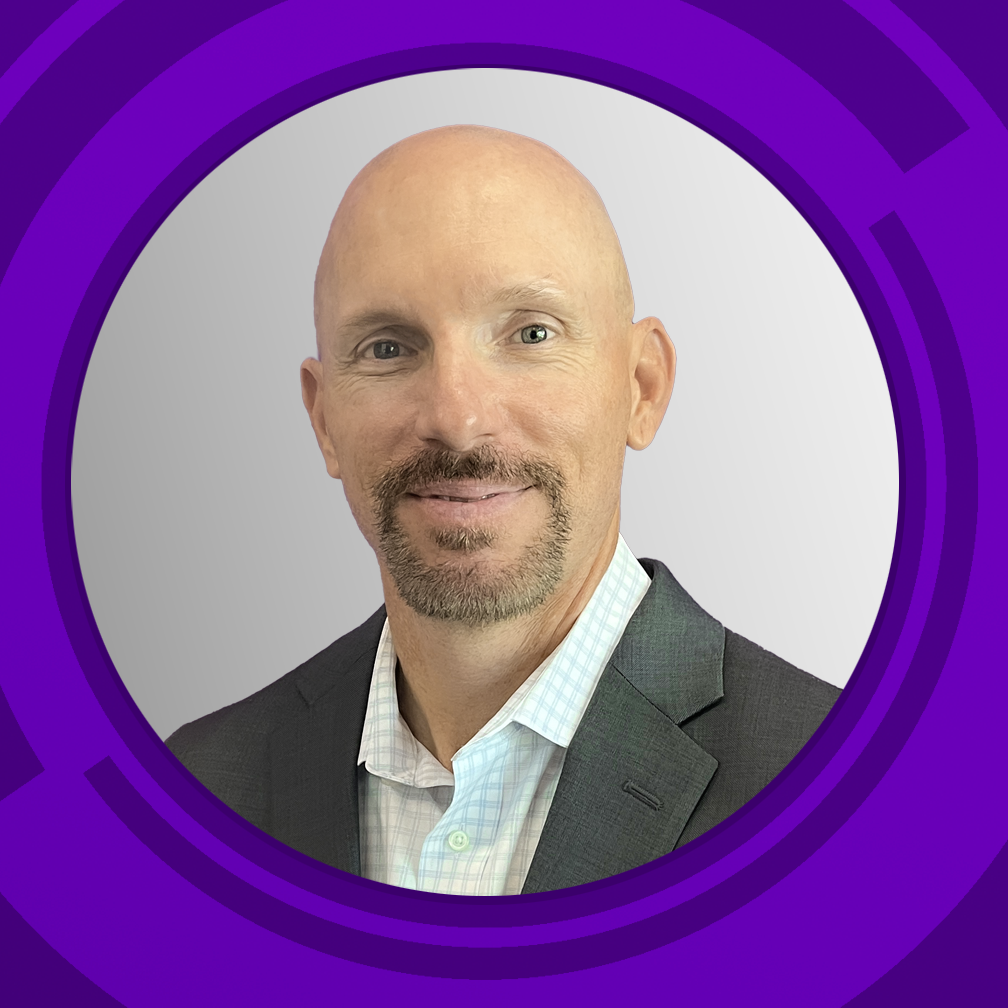 Headshot of a bald man with a goatee, wearing a dark suit jacket and a white button-up shirt, smiling against a plain light gray background, framed by a purple geometric design.