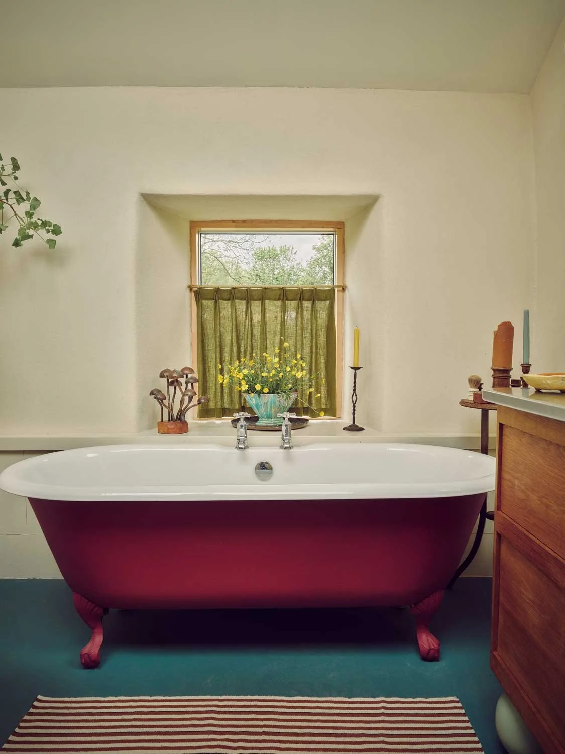 Rustic bathroom designed by Coal Cornwall, featuring a deep crimson red clawfoot roll-top bath, teal painted floorboards, a striped runner rug, and a deep-set window styled with green cafe curtains and fresh flowers.