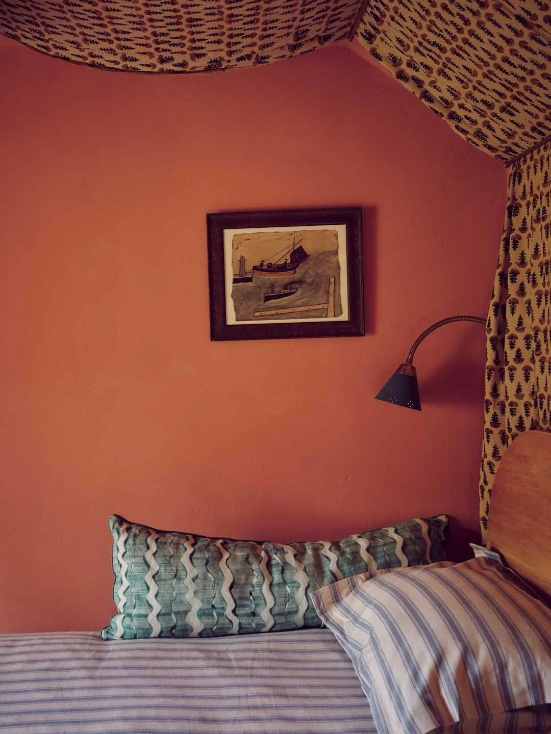 Cornish architectural and interior design by Coal Cornwall showcasing a coastal bedroom with a warm coral wall, framed boat and lighthouse painting, black wall-mounted reading lamp, striped pillow, and pinecone-patterned curtains.