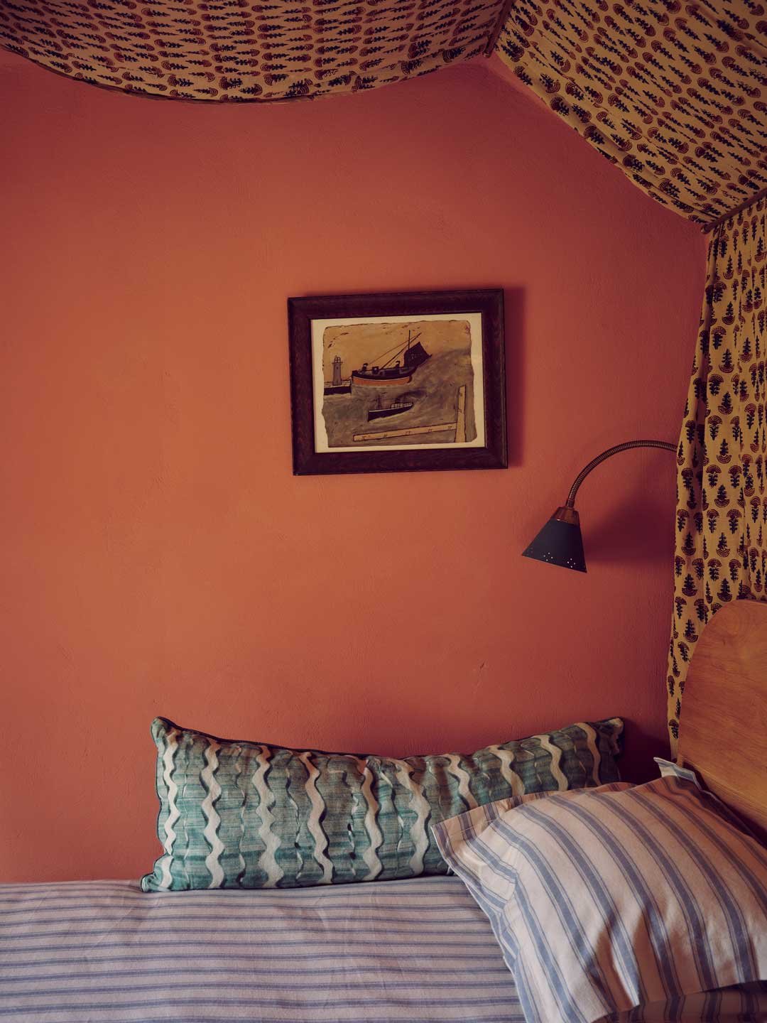 Cohesive coastal bedroom design in Cornwall by Coal Cornwall. Features warm, textured coral pink walls, a framed painting of a boat near a lighthouse, and patterned fabrics including a block-print canopy ceiling and wave-patterned bolster pillow.