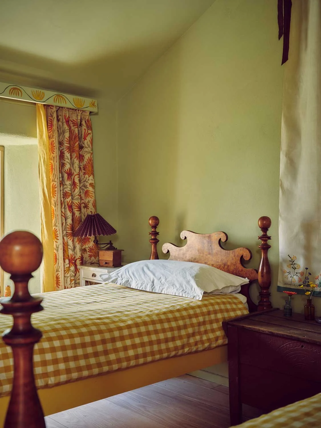 Rustic cottage bedroom by Coal Cornwall featuring sage green plaster walls, a wooden single bed with yellow gingham bedding, hand-painted wooden pelmet with floral curtains, and vintage nightstand styling.