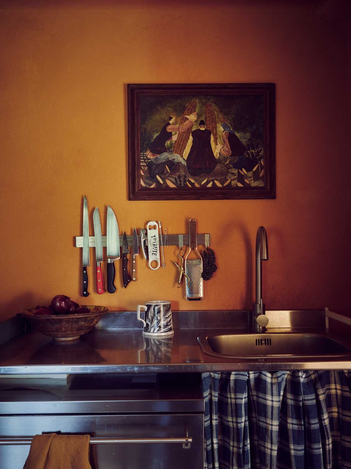Rustic kitchen prep area designed by Coal Cornwall, featuring warm terracotta-toned plaster walls, a stainless steel countertop and sink, a magnetic strip with chef knives, a dark blue plaid fabric sink skirt, and classical framed wall art.