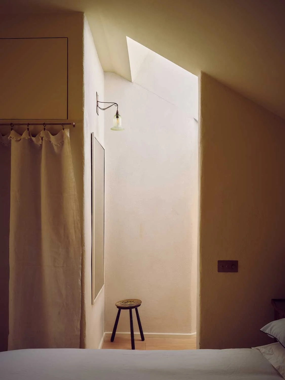 Minimalist hallway interior by Coal Cornwall featuring a high skylight casting natural light onto lime-washed plaster walls, a three-legged wooden stool, a full-length wall mirror, and a delicate wall sconce.