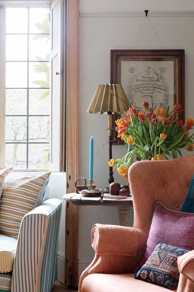 Sunlit living room corner styled by Coal Cornwall, featuring a rustic side table with a pleated brass lamp, a vase of orange tulips, a terracotta wingback armchair, and striped cushions by leaded windows.
