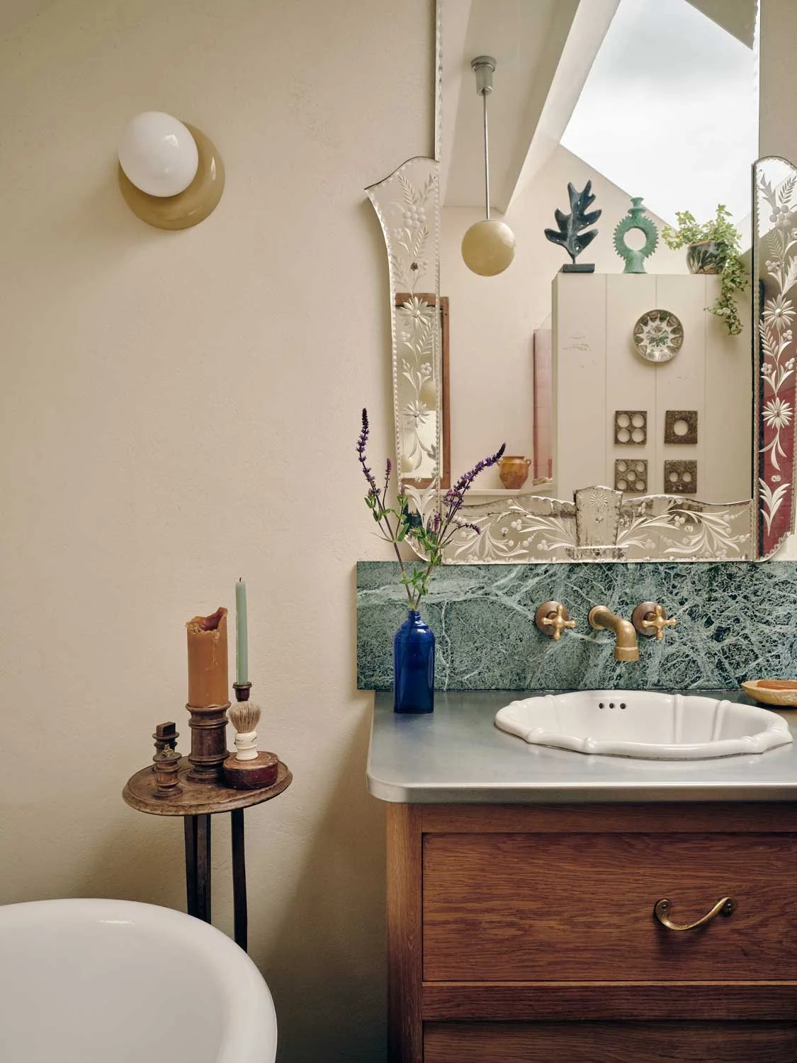 Eclectic and vintage-inspired Cornish bathroom design by Coal Cornwall. Highlights include a detailed, engraved Venetian mirror, a deep green marble backsplash and countertop, a white scalloped sink with brass faucets, and a rustic wooden vanity.