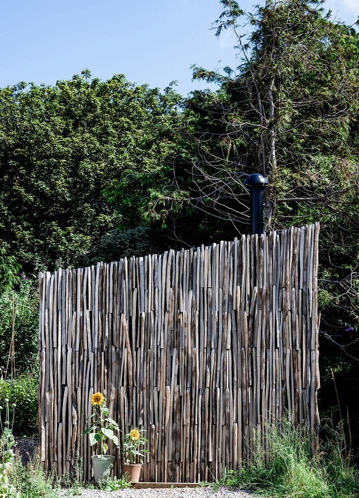 Architectural detail by Coal Cornwall showing a natural split-timber slat facade, a black flue pipe, dense green woodland backdrop, and potted sunflowers along the base.