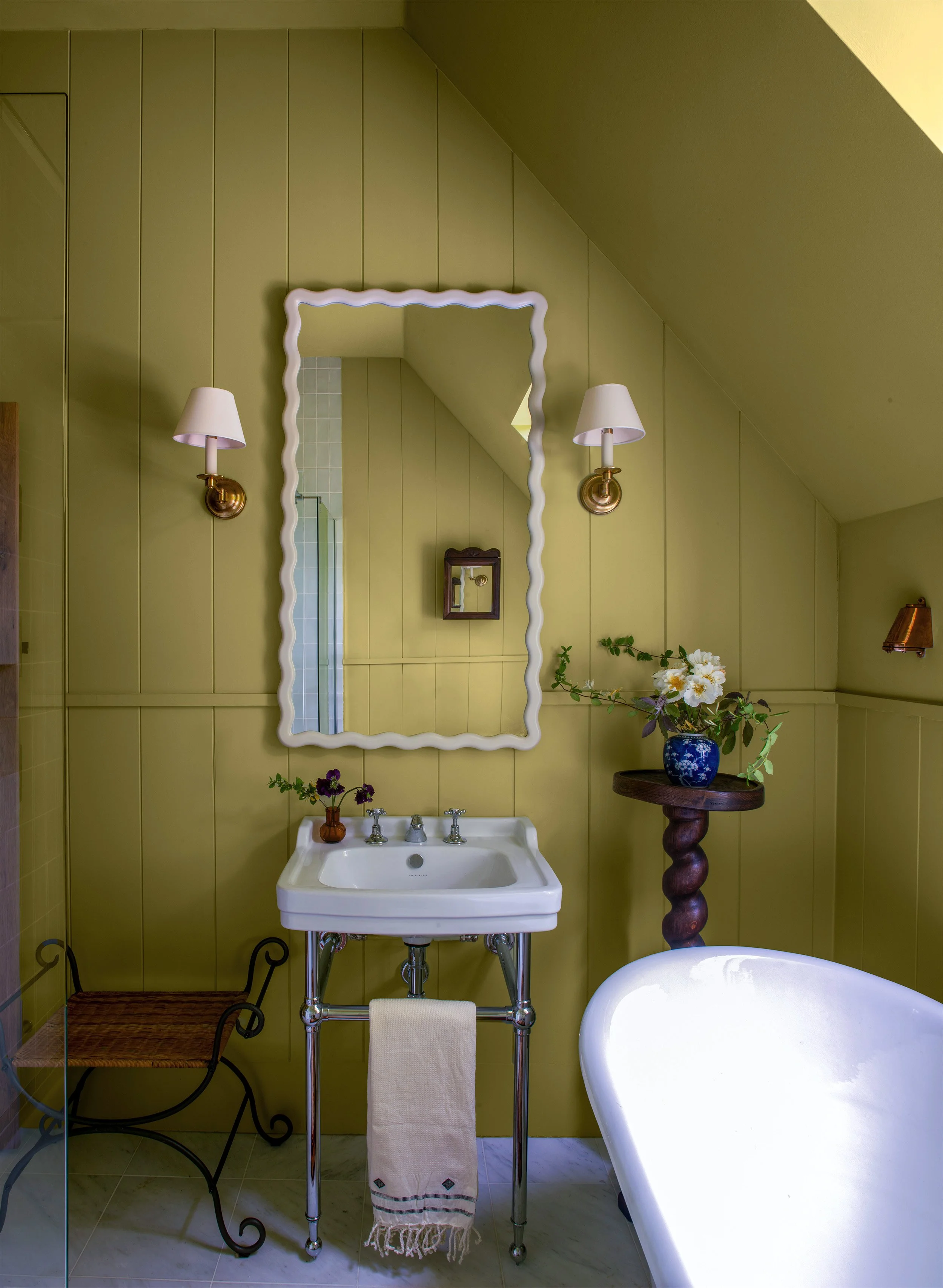 Traditional bathroom interior designed by Coal Cornwall, featuring muted olive green panelling, a scalloped white wall mirror flanked by brass sconces, a chrome console basin, and a freestanding tub.