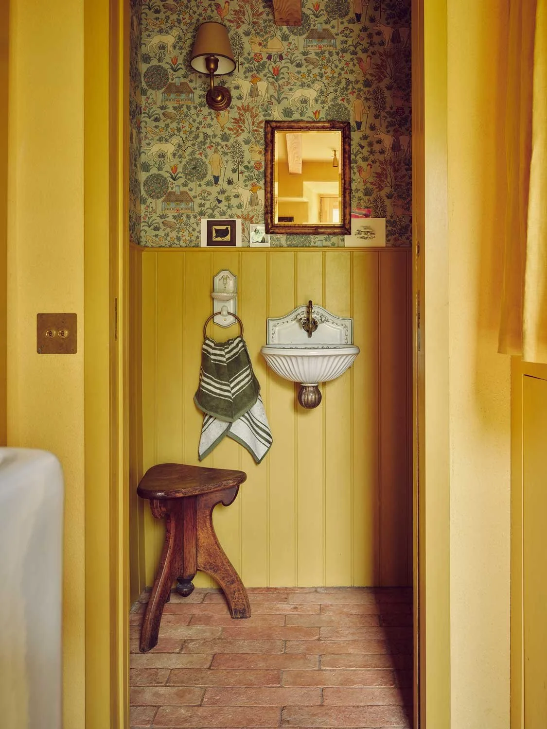 Charming powder room designed by Coal Cornwall, featuring yellow tongue-and-groove wall panelling, patterned wallpaper, a wall-mounted scalloped shell basin, rustic wooden stool, and terracotta brick flooring.