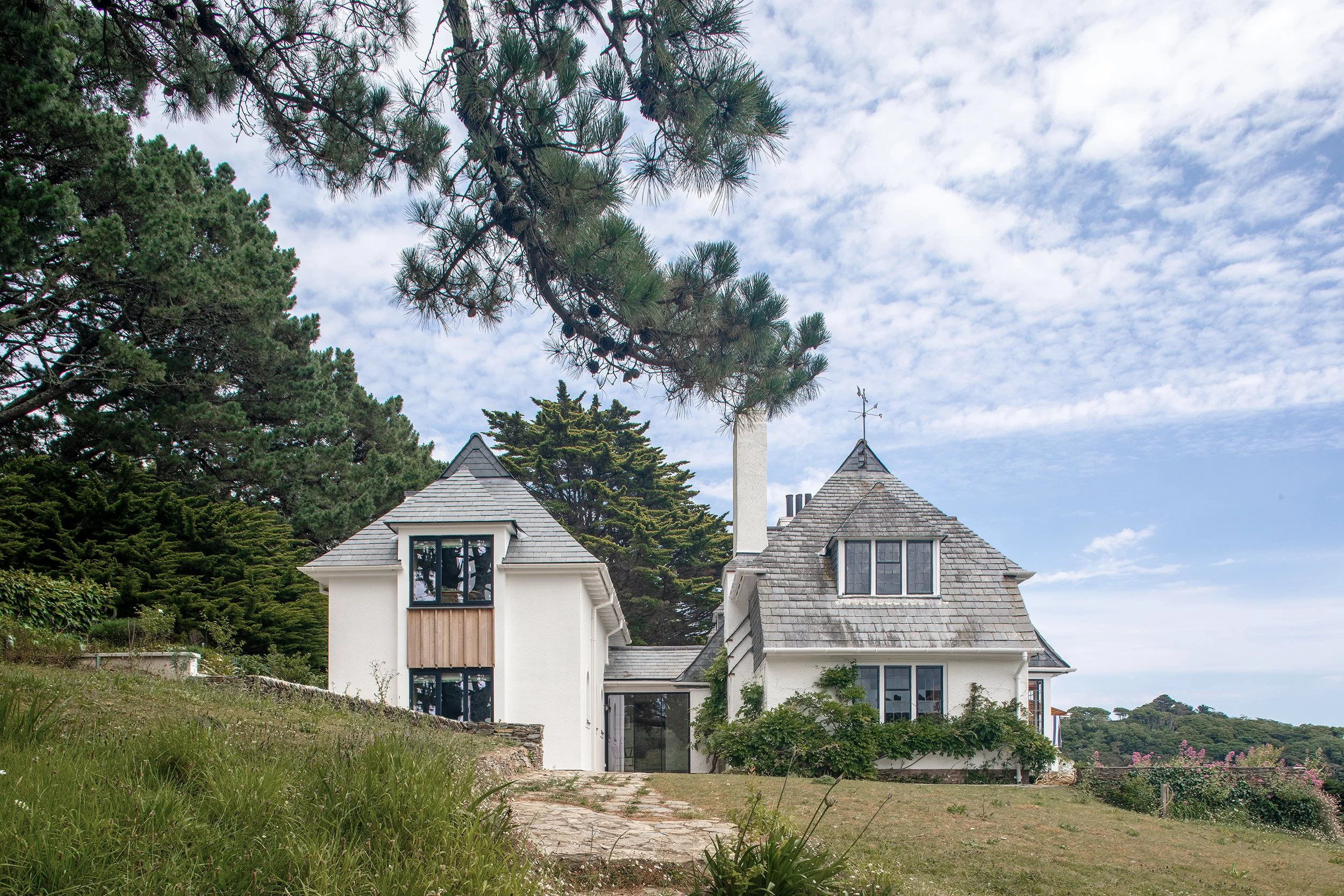 Coastal house extension designed by Coal Cornwall, featuring pitched slate roofs, crisp white render, timber infill cladding panels, a tall chimney stack, and mature pine trees set on a grassy hill.