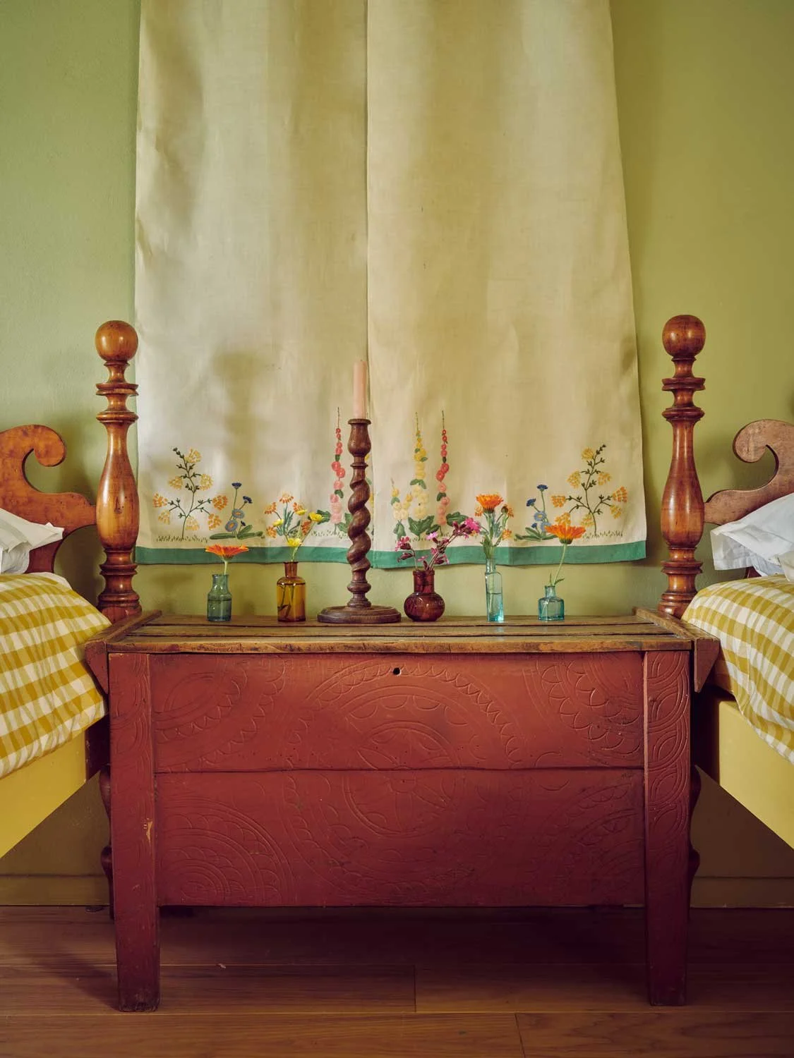 Twin cottage bedroom designed by Coal Cornwall, featuring an antique carved red-painted wooden chest between two wooden beds, styled with botanical curtain fabric, small glass flower vases, and a turned wooden candlestick.