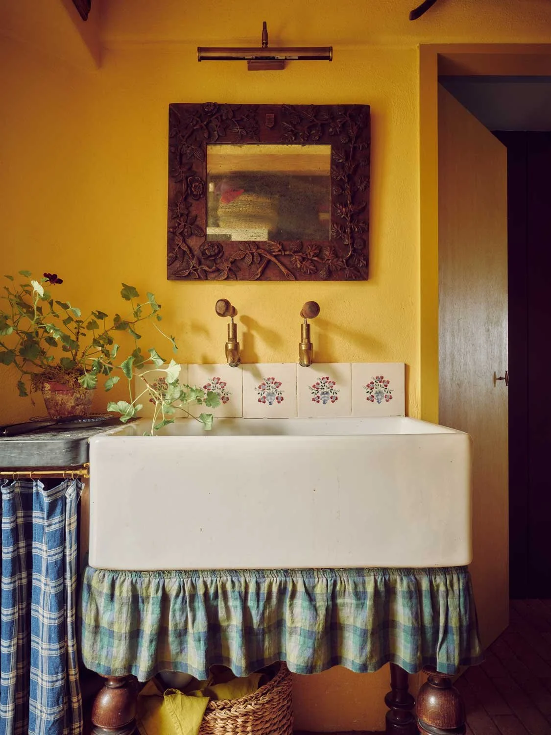 Rustic utility room by Coal Cornwall featuring a deep white Belfast sink styled with a green-and-yellow plaid fabric skirt, vintage brass taps, hand-painted floral tiles, and vibrant yellow walls with an carved wood mirror.