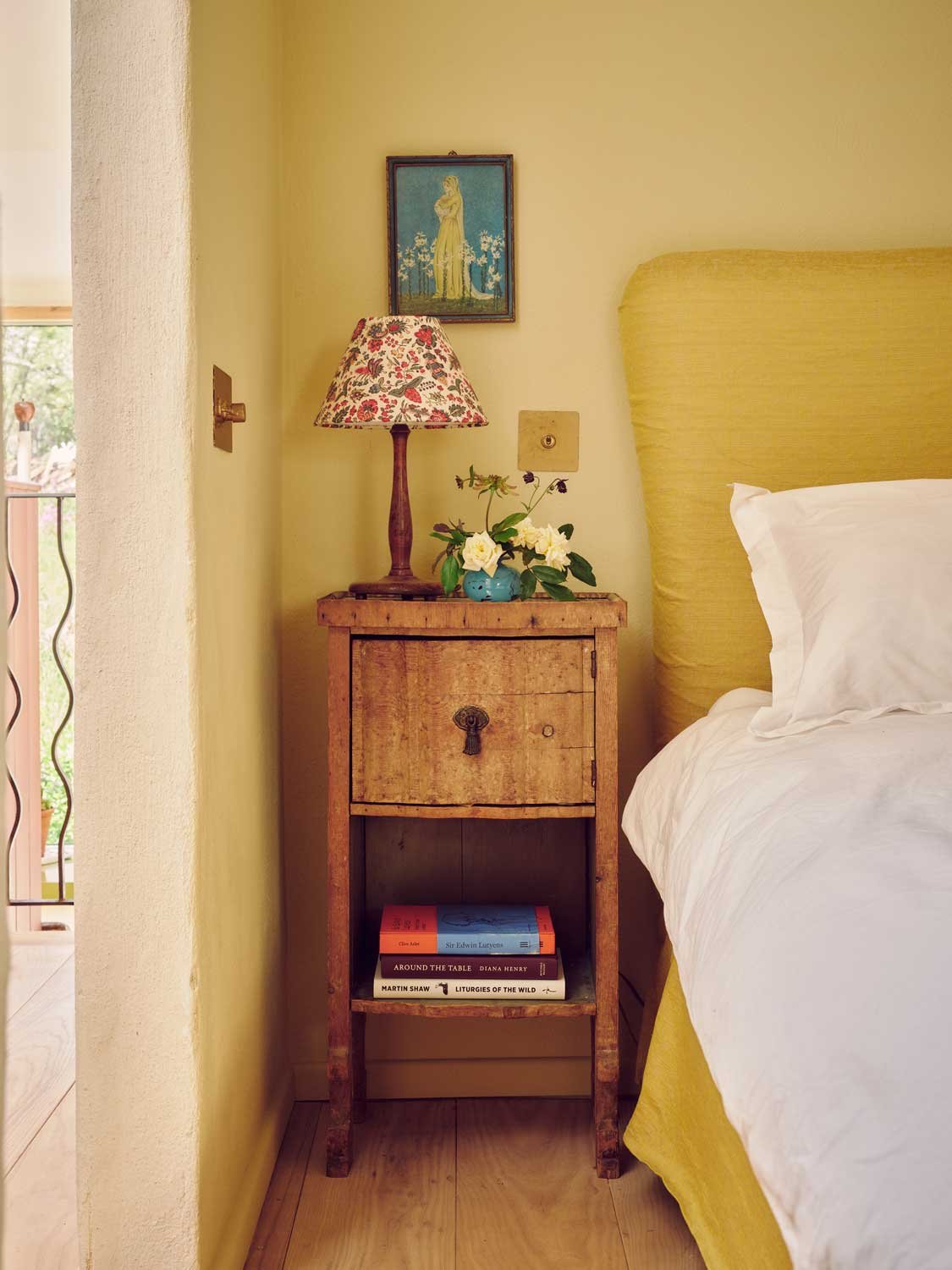 Cozy bedside setup designed by Coal Cornwall, featuring a vibrant yellow fabric headboard, a weathered rustic wooden nightstand with a floral lampshade, and warm lime-washed walls.