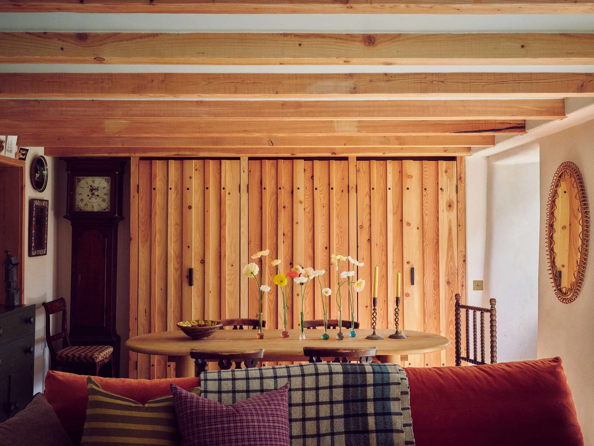 Bespoke dining room design by Coal Cornwall featuring a floor-to-ceiling slatted timber wall, exposed ceiling beams, a vintage grandfather clock, and an oval wooden table decorated with delicate flower vases.
