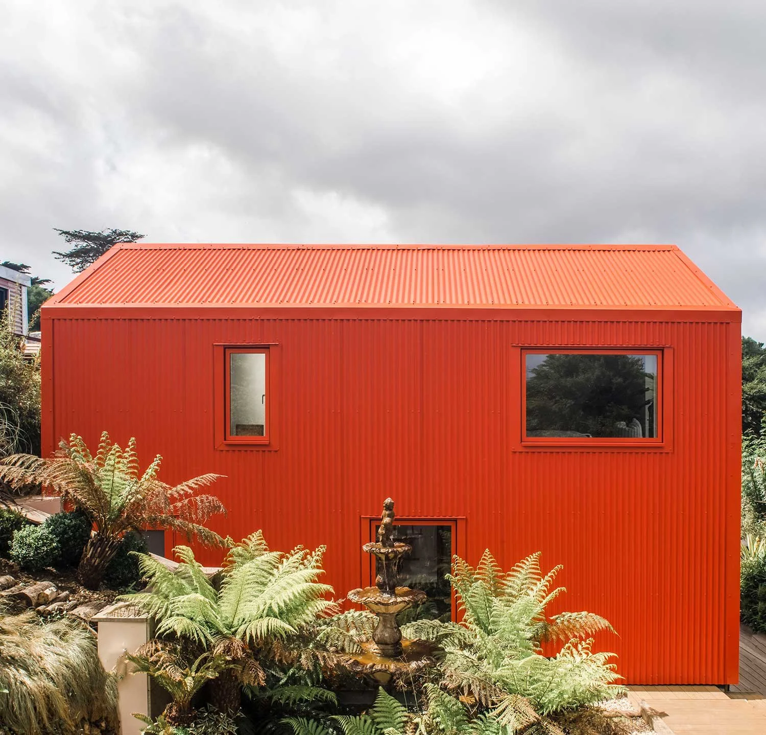 Modern barn exterior designed by Coal Cornwall, featuring vibrant orange-red corrugated metal cladding, matching window frames, lush tree ferns, and a classic tiered stone water fountain in the foreground garden.