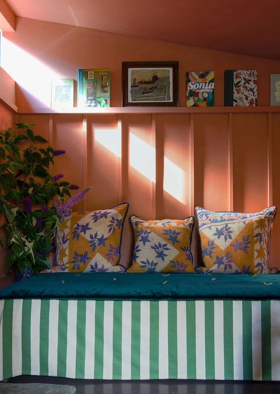 Warm terracotta-painted studio interior by Coal Cornwall, featuring tongue-and-groove wall panelling, a picture ledge with art books, and a green-striped fabric daybed with pattern-matched cushions.