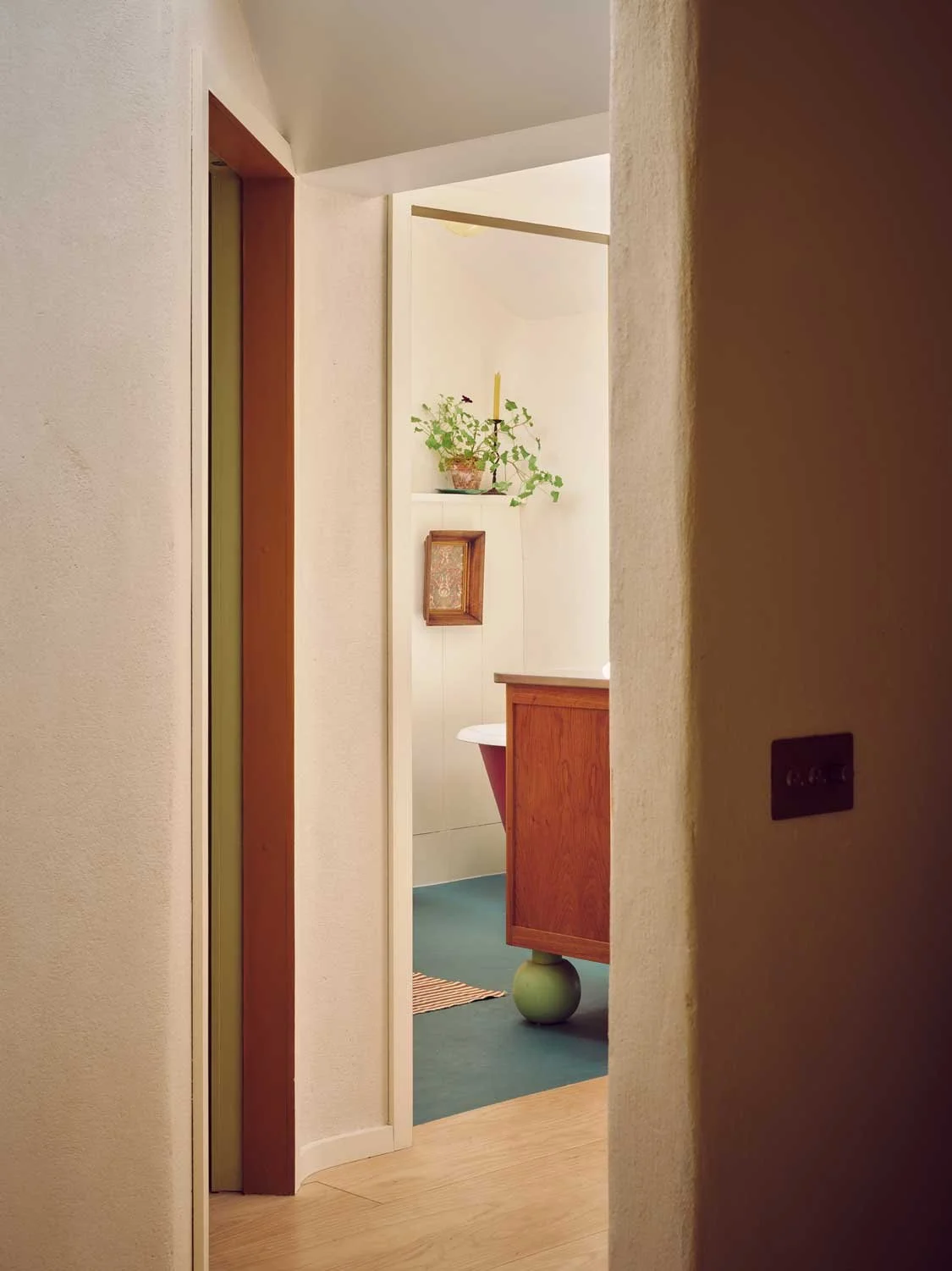 Architectural doorway perspective by Coal Cornwall, looking into a bathroom with a bespoke wooden vanity resting on a green spherical foot, teal painted flooring, and lime-washed plaster walls.