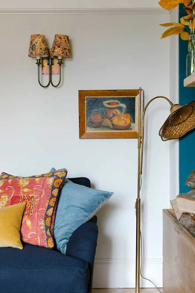 Detailed reading nook styled by Coal Cornwall, featuring patterned fabric wall sconces, a still life oil painting, a brass floor lamp with a woven shade, and vibrant throw cushions on a navy sofa.