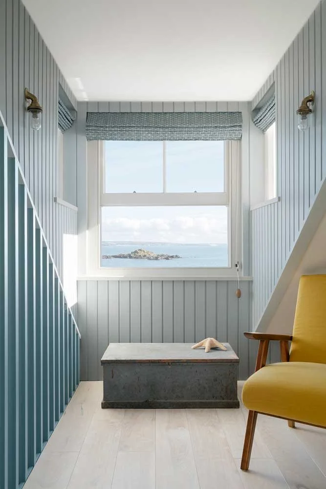 Coastal hallway landing designed by Coal Cornwall, featuring pale blue tongue-and-groove panelling, a sea view sash window with patterned blinds, a rustic wooden chest, and a mustard yellow armchair.