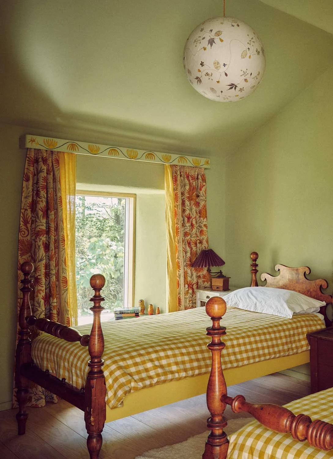 Charming children's bedroom designed by Coal Cornwall, featuring muted sage green walls, antique four-poster wooden single beds with yellow gingham bedding, floral curtains, and a botanical paper globe light fixture.
