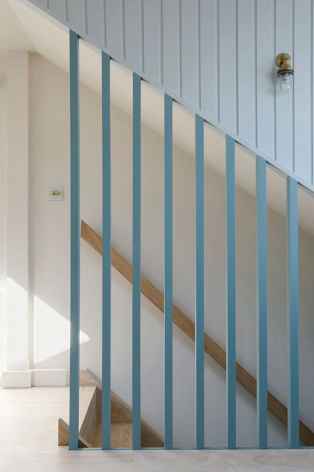 Architectural interior by Coal Cornwall featuring a modern coastal staircase with slate-blue vertical slat balusters, light timber treads, pale blue vertical wall panelling, and brass nautical wall lights.