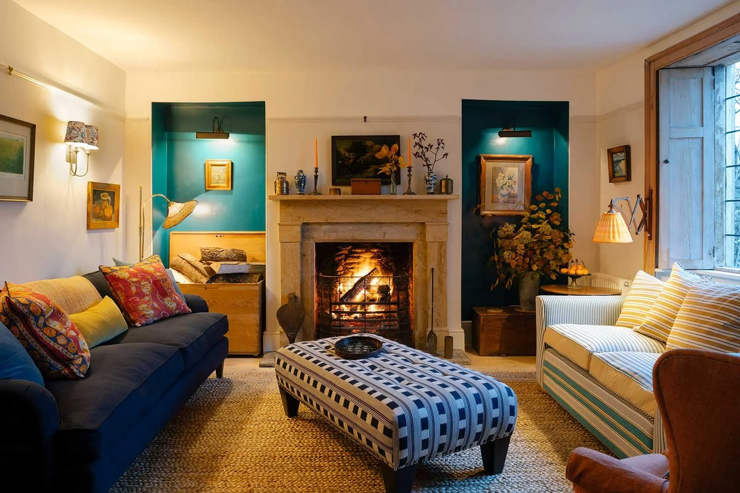 Cosy living room designed by Coal Cornwall, featuring an open stone fireplace, deep teal painted alcoves, a blue sofa, a geometric pattern ottoman, and a striped loveseat by a window shutter.