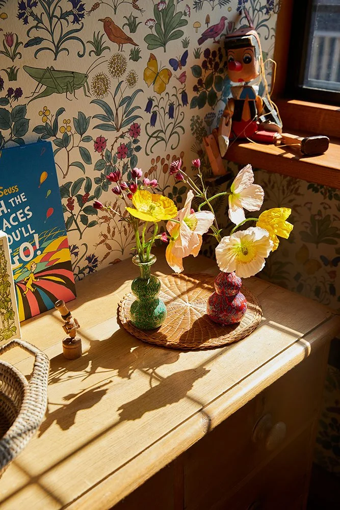 Charming nursery interior styled by Coal Cornwall, featuring botanical insect wallpaper, delicate poppies in mini bud vases on a pine dresser, Dr. Seuss book, and wooden Pinocchio figurine.