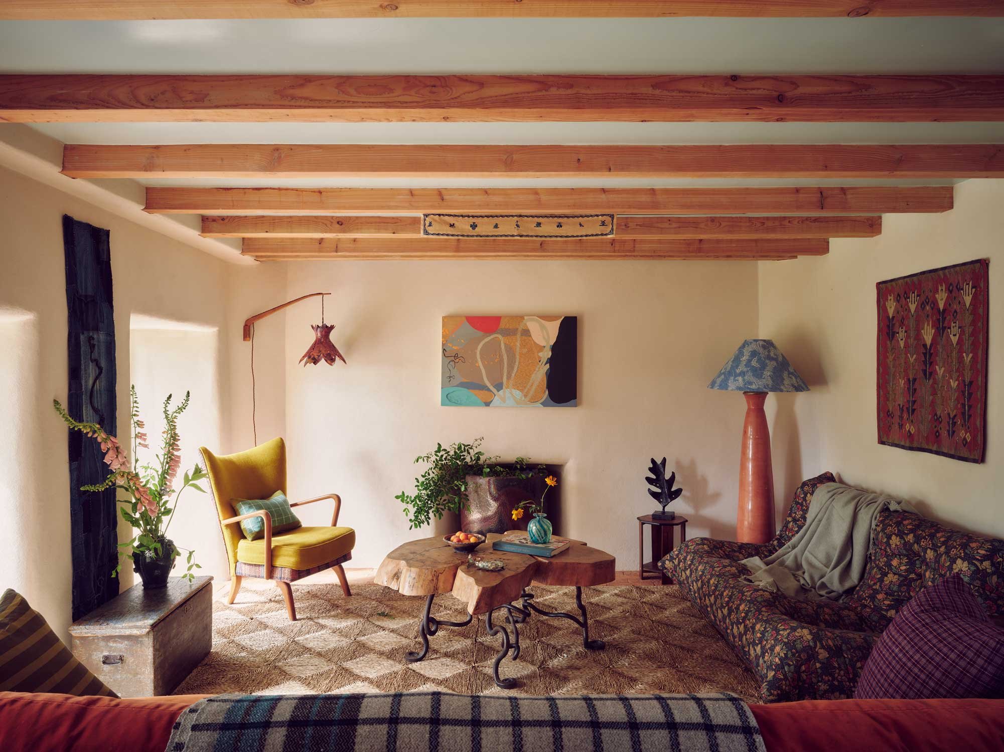 Rustic living room design by Coal Cornwall, featuring exposed wooden ceiling beams, a mustard-yellow wingback chair, live-edge coffee table, patterned sofa, and textured plaster walls adorned with modern art and textiles.