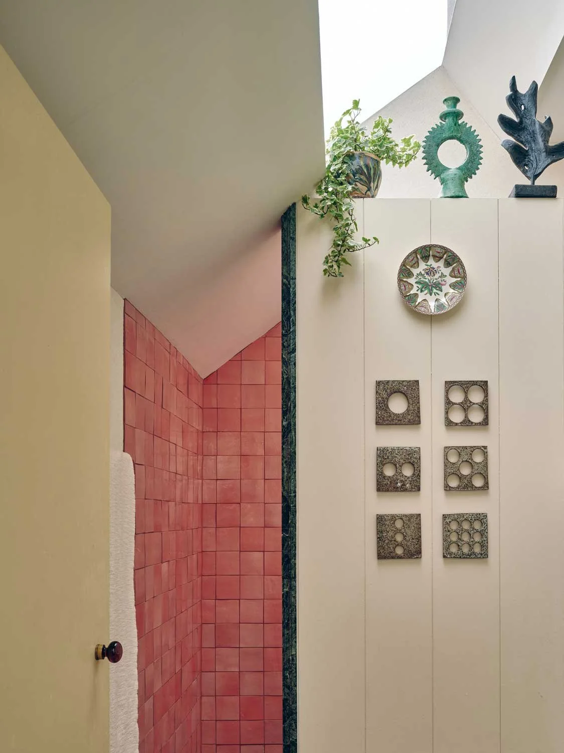 Unique Cornish bathroom designed by Coal Cornwall, featuring high-gloss coral pink square wall tiles contrasted with a marble corner border, decorative plates, geometric wall art, and an overhead skylight.
